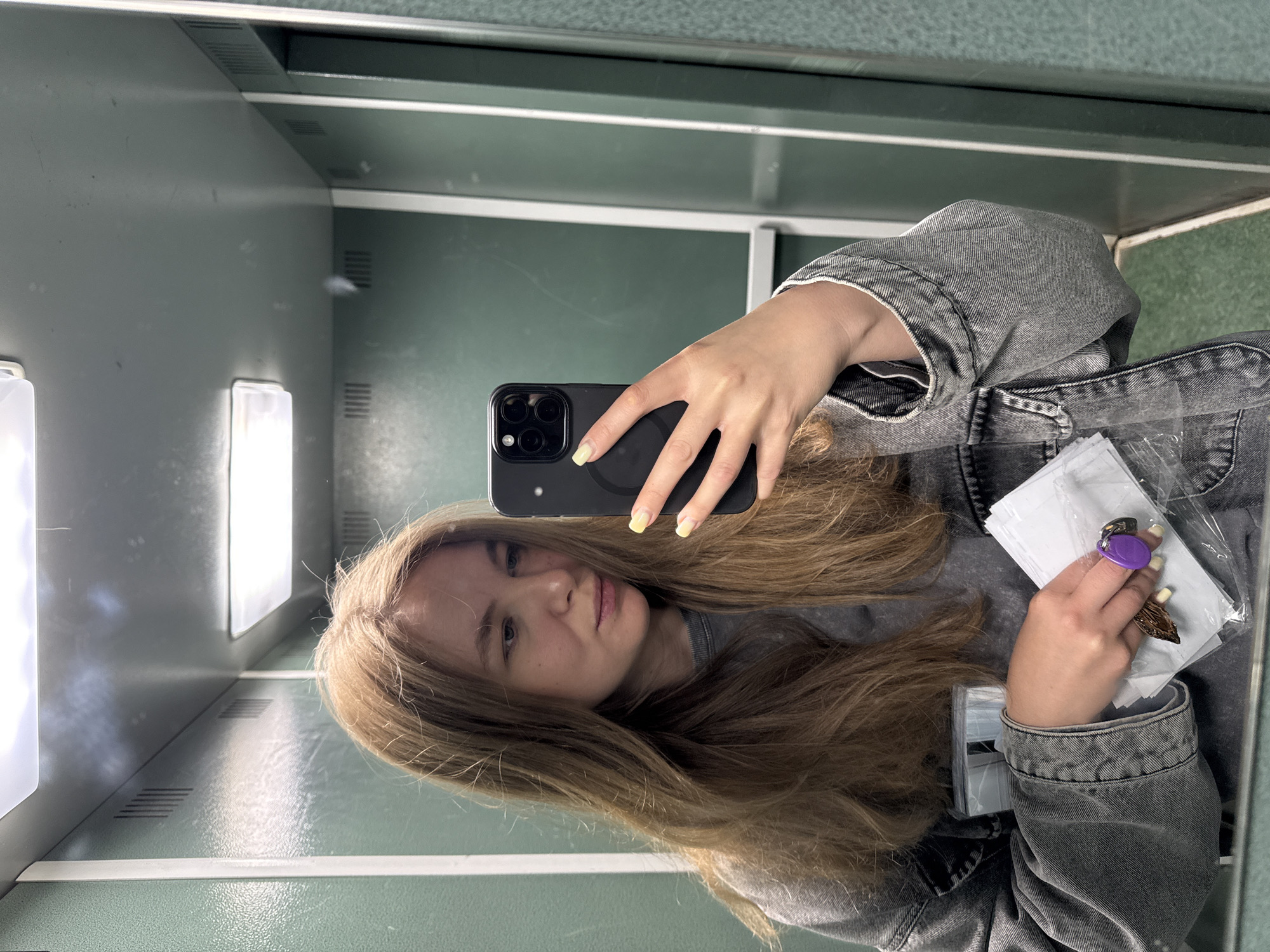 A fair-skinned woman with long, light brown hair is taking a selfie in a location that appears to be an elevator. She is wearing a gray denim jacket and a gray shirt. The woman is holding a black smartphone in her right hand, displaying the rear camera. Her left hand holds a small, wrapped item with a purple, rounded object on top. The interior of the elevator has a gray-green color scheme, with horizontal lines indicating panels or surfaces. The lighting is artificial, with visible light fixtures reflected in the elevator's walls. The location is an elevator, but the specific city in Russia is not discernible from the image.