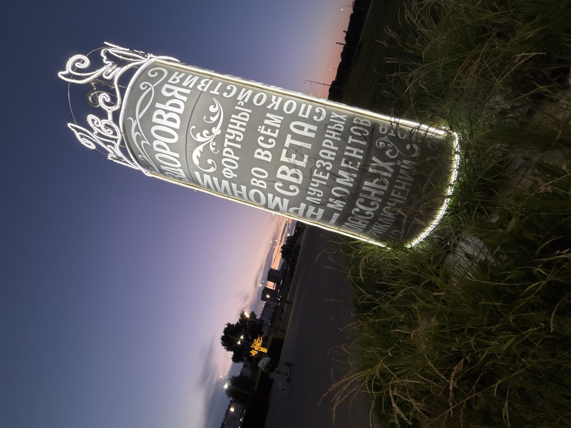 This media file shows an illuminated cylindrical object with text inscribed on it, set against a twilight sky and surrounded by tall grass. The object appears to be a sign or monument, featuring ornate white lettering against a dark gray background. The text is in Russian and includes words like "ЗДОРОВЬЯ" (health), "СЧАСТЬЯ" (happiness), "СВЕТА" (light), "МОМЕНТОВ" (moments), and "УЧЕЖЕНИЙ" (teachings).
The sky is a deep purple and blue, suggesting the time of day is dusk or early evening. There are faint hints of orange and pink near the horizon, indicating a sunset. In the background, there are silhouetted buildings and streetlights, suggesting an urban or suburban environment. The grass is tall and appears wild, with some blades illuminated by the light from the object.
The overall mood is serene and slightly mysterious, due to the twilight lighting and the illuminated, text-covered object. The focus is on the sign and its messages, which seem to convey positive aspirations. The composition is slightly tilted, adding a dynamic feel to the scene. There are no visible people in the image. The weather appears clear and calm.