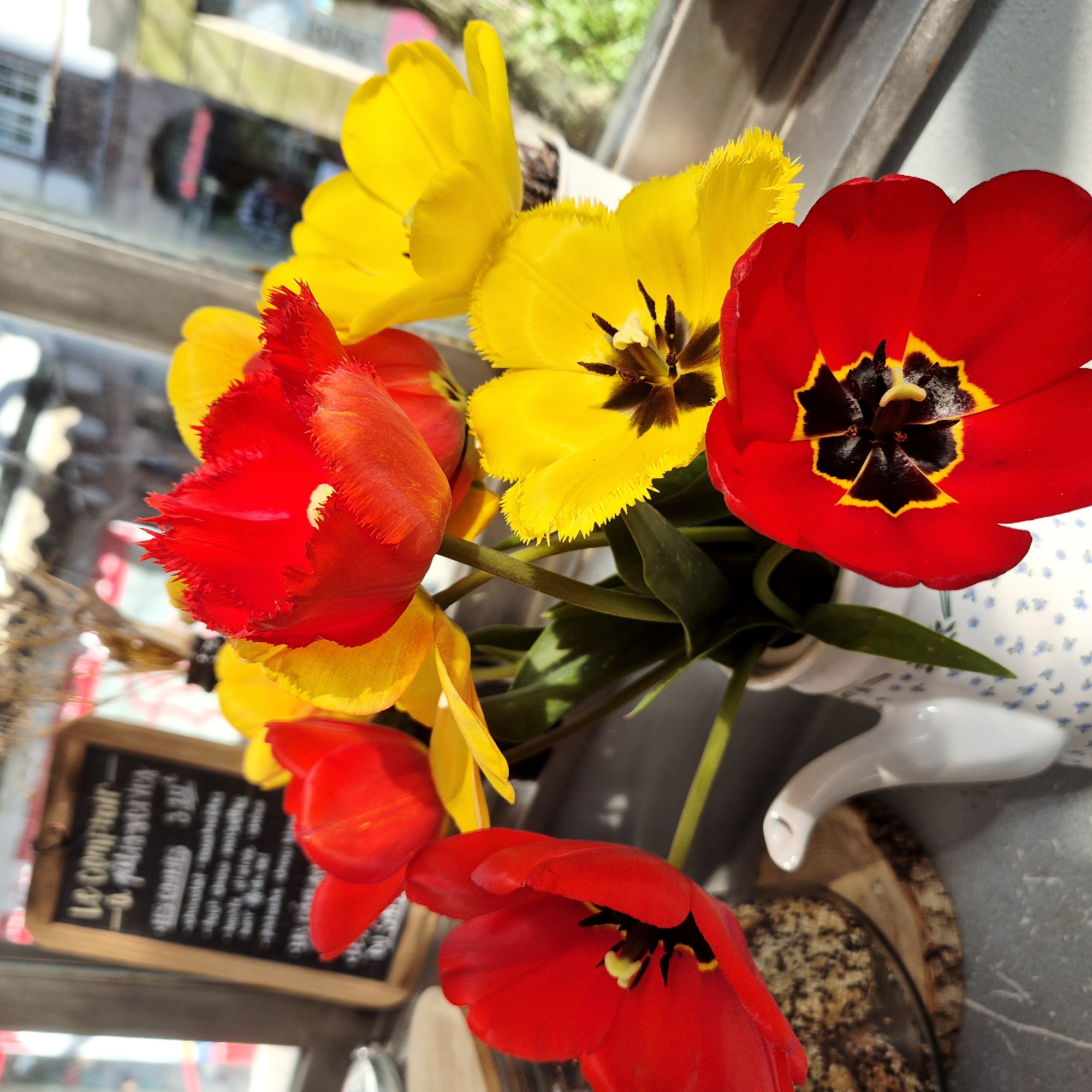 A still-life photograph taken in Strasbourg, France, depicts an arrangement of red and yellow tulips in a small, white porcelain vase with a blue floral pattern. The vase sits on a wooden slice, which itself rests on a grey surface near a window.
A chalkboard menu, with the words "Le Comptoir a" and some written prices, partially obscured by the flowers, leans against the window. The tulips are in full bloom, showing the interior details of their petals.
Through the window, a blurred urban street scene is visible, with buildings and possibly people. The lighting suggests it is daytime.