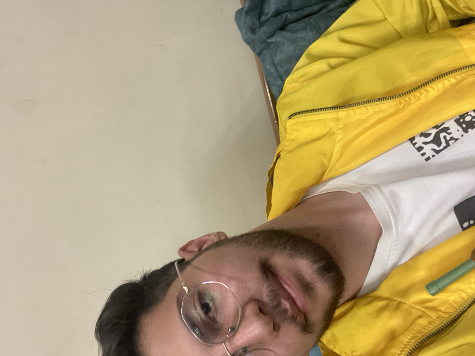 The image displays a head and upper torso shot of a young adult male in an indoor setting. The male subject has dark, short hair, a trimmed beard, and a mustache. He wears round, thin metal-rimmed glasses and looks directly at the camera with a slight smile. He is dressed in a bright yellow zippered jacket over a white t-shirt that features a partial black and white graphic design. A small, light green cylindrical object, possibly a plastic straw or similar item, is visible in the lower left portion of the frame, held near his chest. The background consists of a plain, light-colored wall, with a horizontal wooden edge and a folded blue-green fabric visible in the upper right. The specific location within Homs, Syria, cannot be determined from the visual information presented.