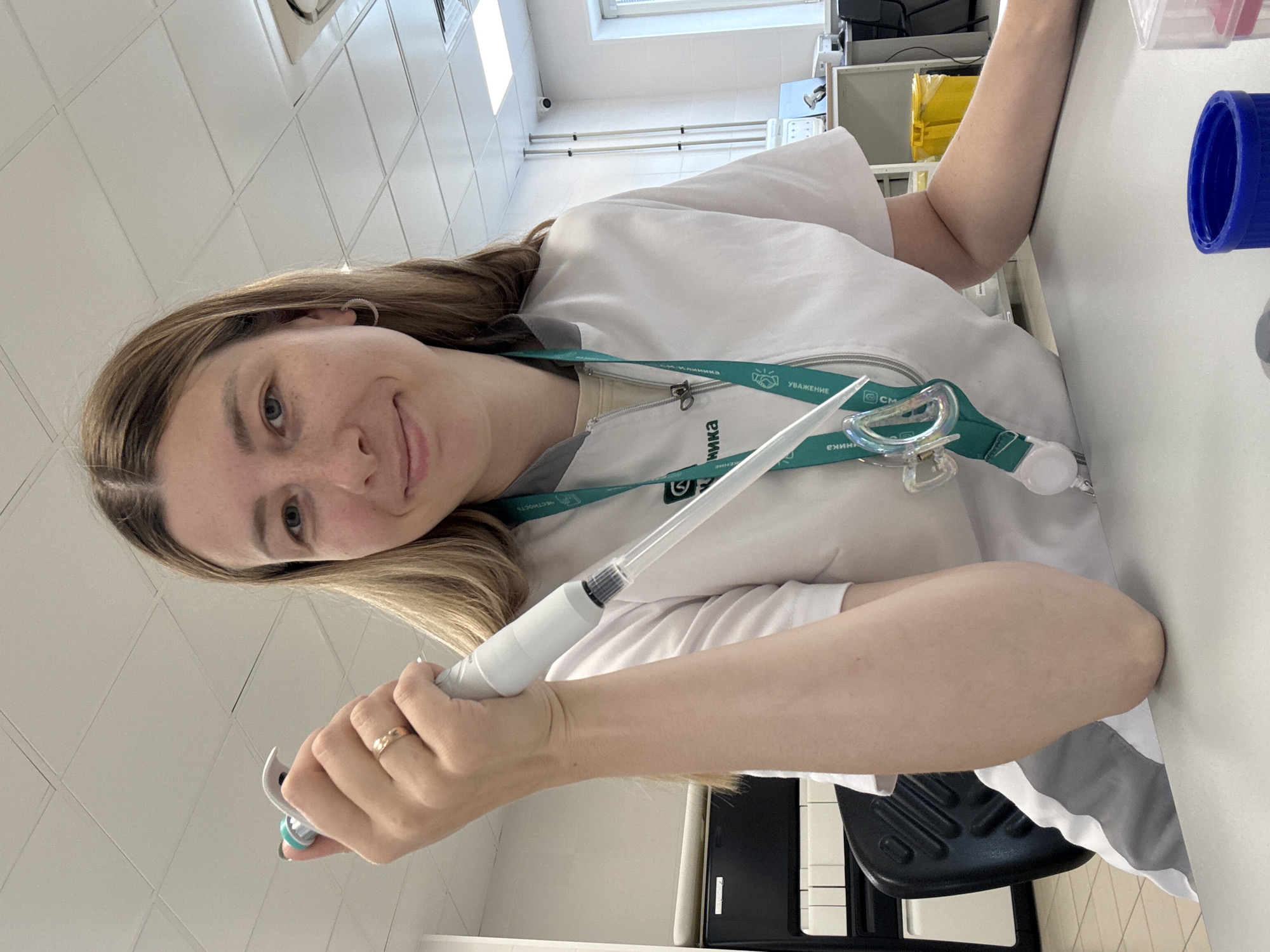 A woman in a white lab coat is holding a pipette. She is wearing a green lanyard with a badge that says "Клиника" and "УВАЖЕНИЕ", indicating she is likely at a medical clinic. The setting appears to be a laboratory or clinic room, with white walls and a white countertop. Behind her, there's a glimpse of a cabinet with yellow containers. The exact location within Moscow, Russia, cannot be determined from the image alone.