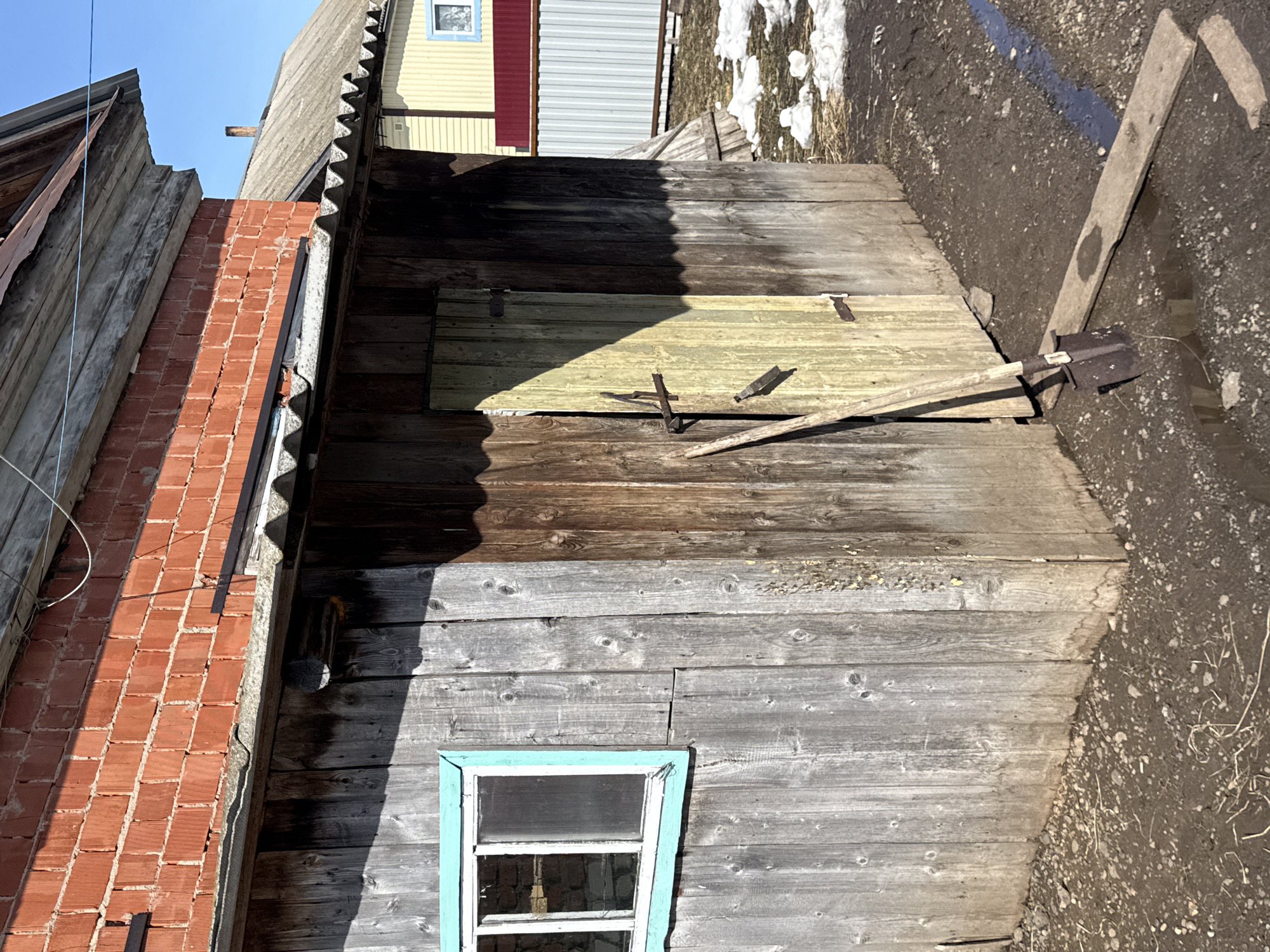 The image presents an external view of an old, weathered wooden structure, possibly a shed or outbuilding, located in what is presumed to be Кипчак, Russia. The structure features a small window with a light blue frame. To the left of the wooden structure, there is a red brick chimney or wall. The roof is corrugated and made of gray metal. The wall is constructed of old wooden planks of different colors, which were exposed to the sun and weather. A shovel is leaning against the exterior, a section of which has been painted yellow. The ground surrounding the structure is dirt with bits of debris scattered around. There is snow and ice in the background along with a building in the distance that is painted yellow and red. The sky is clear and light blue.