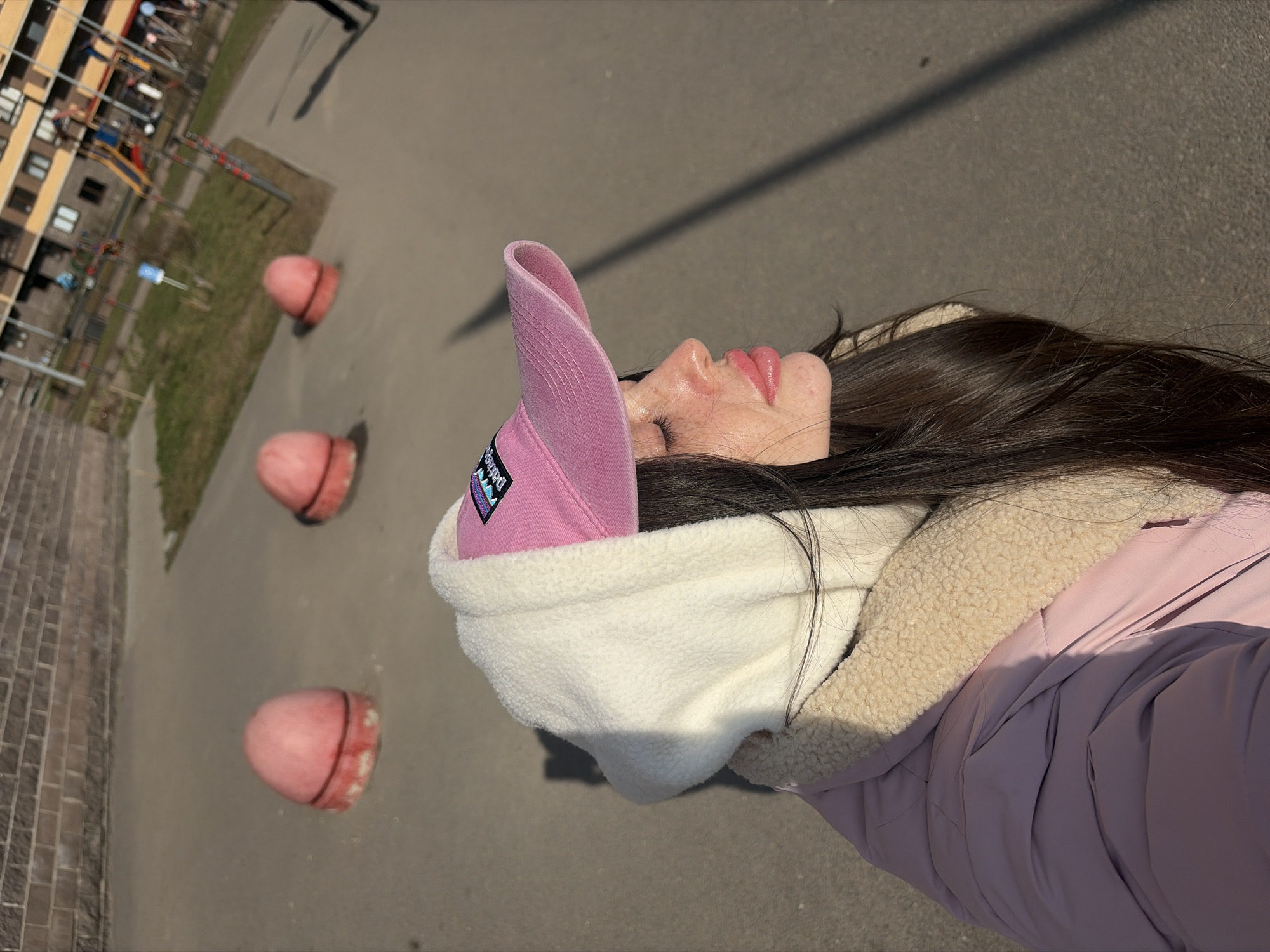 A top-down, medium-shot photograph depicts a woman on a sunny day in what appears to be Saint Petersburg, Russia.
The woman is wearing a pink visor with a visible logo, a white fleece hood, and a light purple winter jacket. Her dark hair is visible extending from under the hood. She is lying on her back.
The background shows an outdoor public space with three red spherical objects spaced out in a line on the ground. Adjacent to the spheres is a brick pathway and a patch of grass that runs alongside a paved area. In the distance, buildings and what looks like a playground are visible. The shadow cast across the pavement suggests that the picture was taken in a well-lit environment.