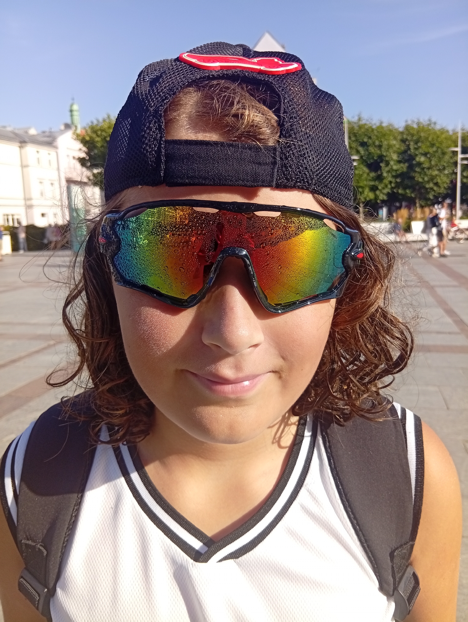 A young person, facing forward, is pictured outdoors in Sopot, Poland. The individual is wearing a black baseball cap with a red accent, mirrored sunglasses with a rainbow tint and water droplets, and a white athletic tank top with black trim. Their hair is brown and curly, falling around their face and shoulders. The background shows a street scene with buildings, trees, and a clear blue sky. The overall impression is of a sunny day in a public space.