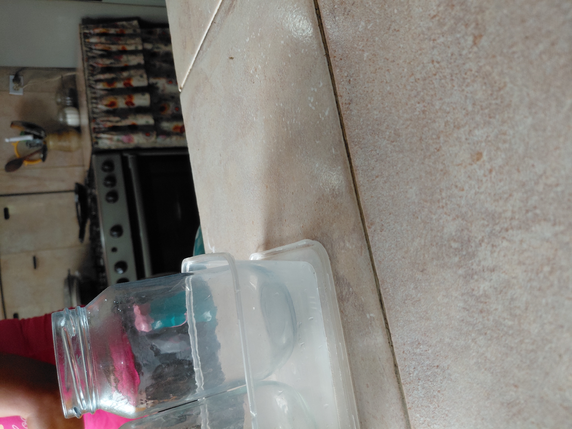 This image, taken in what appears to be a kitchen in Santa Ana, Venezuela, is rotated on its side. In the foreground, a person's arm, wearing a red or pink shirt, is visible as they hold or interact with a large, clear plastic or glass jar. Next to the jar, a clear plastic ice cube tray is present, possibly containing water or ice.
The background reveals a domestic kitchen setting. A light-colored tiled counter or wall extends across much of the right side and top of the image. On a surface further back, a small jar filled with a white substance (likely salt or sugar) and a yellow container holding kitchen utensils (spoons, spatulas) are visible. Below these items, a black stovetop or oven with circular knobs can be seen. Above the stovetop, a patterned fabric, possibly a curtain or towel, hangs on a shelf. A white electrical outlet is also visible on the wall. The lighting suggests it is daytime. No specific activity beyond the person handling the jar is clearly depicted, and no text is visible.