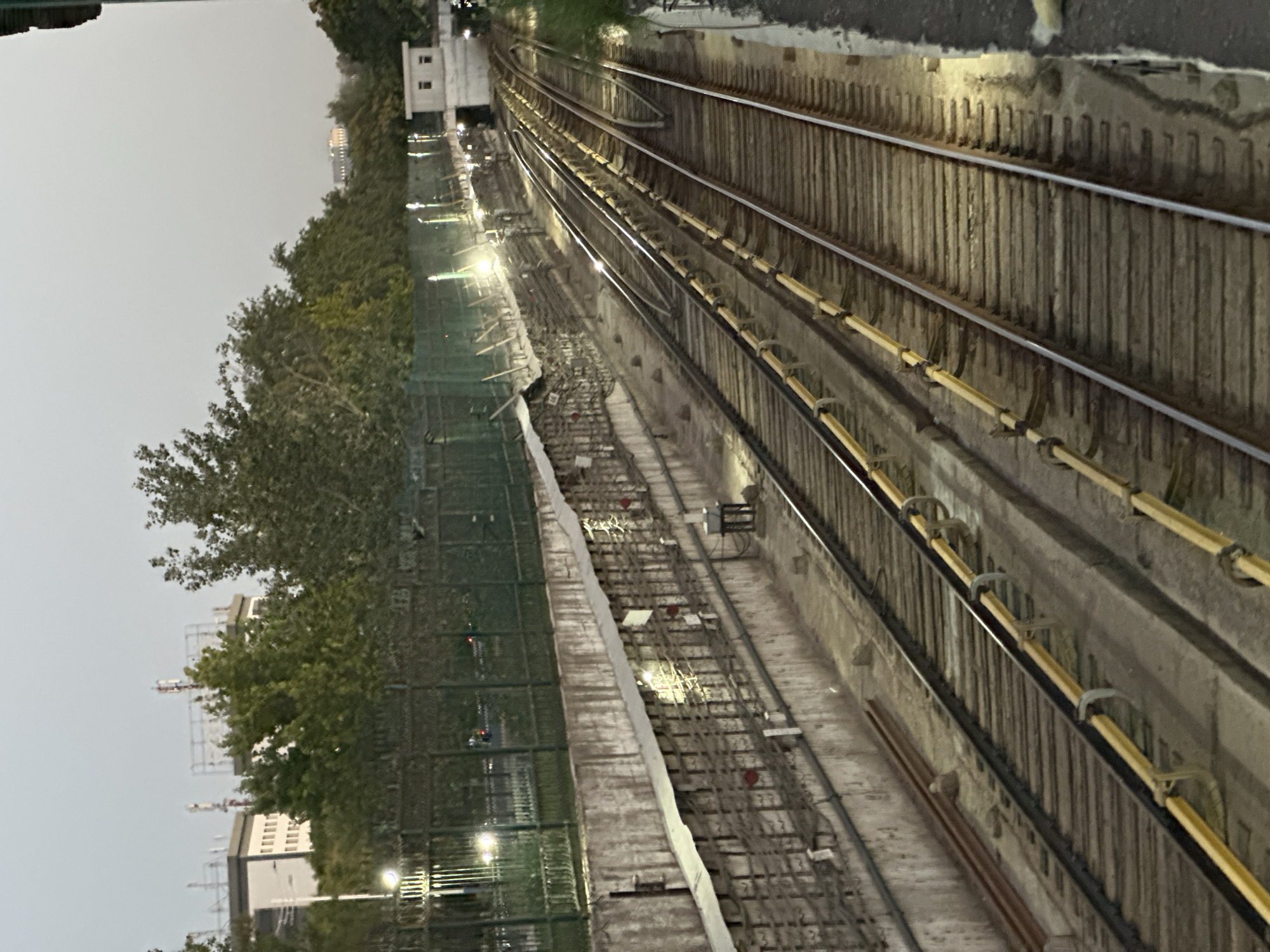 The image depicts a view of railway tracks, likely a metro or train line, in Tashkent, Uzbekistan. The tracks run diagonally across the frame, receding into the distance. The scene is illuminated by artificial lights, suggesting it is either dusk or nighttime. Overhead, power lines and a green fence are visible, bordering the tracks. Buildings and trees are present in the background, with one building in the far distance appearing to be a prominent landmark with illuminated windows. Construction materials, such as metal pipes, are scattered near the tracks. The overall impression is one of an urban infrastructure scene, possibly undergoing maintenance or development.