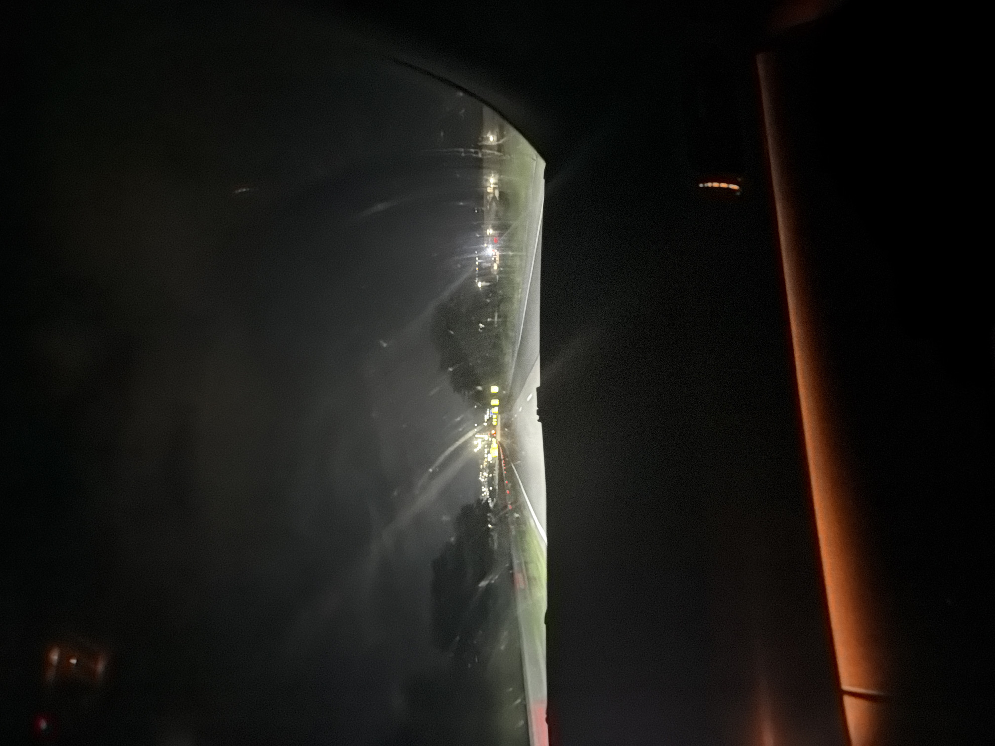 The image presents a nighttime view from within a vehicle, looking forward through a windshield. The scene is situated on a highway or major road in Krepost, Bulgaria. The lower portion of the frame is occupied by the vehicle's dark dashboard, which features a prominent amber-colored ambient light strip. The road ahead is illuminated by the vehicle's headlights, revealing white lane markings. On the left side of the road, faint orange traffic cones or barriers are visible. In the middle distance, clusters of bright yellow and white lights indicate an urban area or significant lighting infrastructure, alongside discernible red taillights from other vehicles. The sky above is dark, suggesting overcast conditions. The windshield appears to have moisture or debris, causing light sources to streak and blur. Blurred reflections of internal lights are visible on the upper section of the windshield. No individuals are visible.