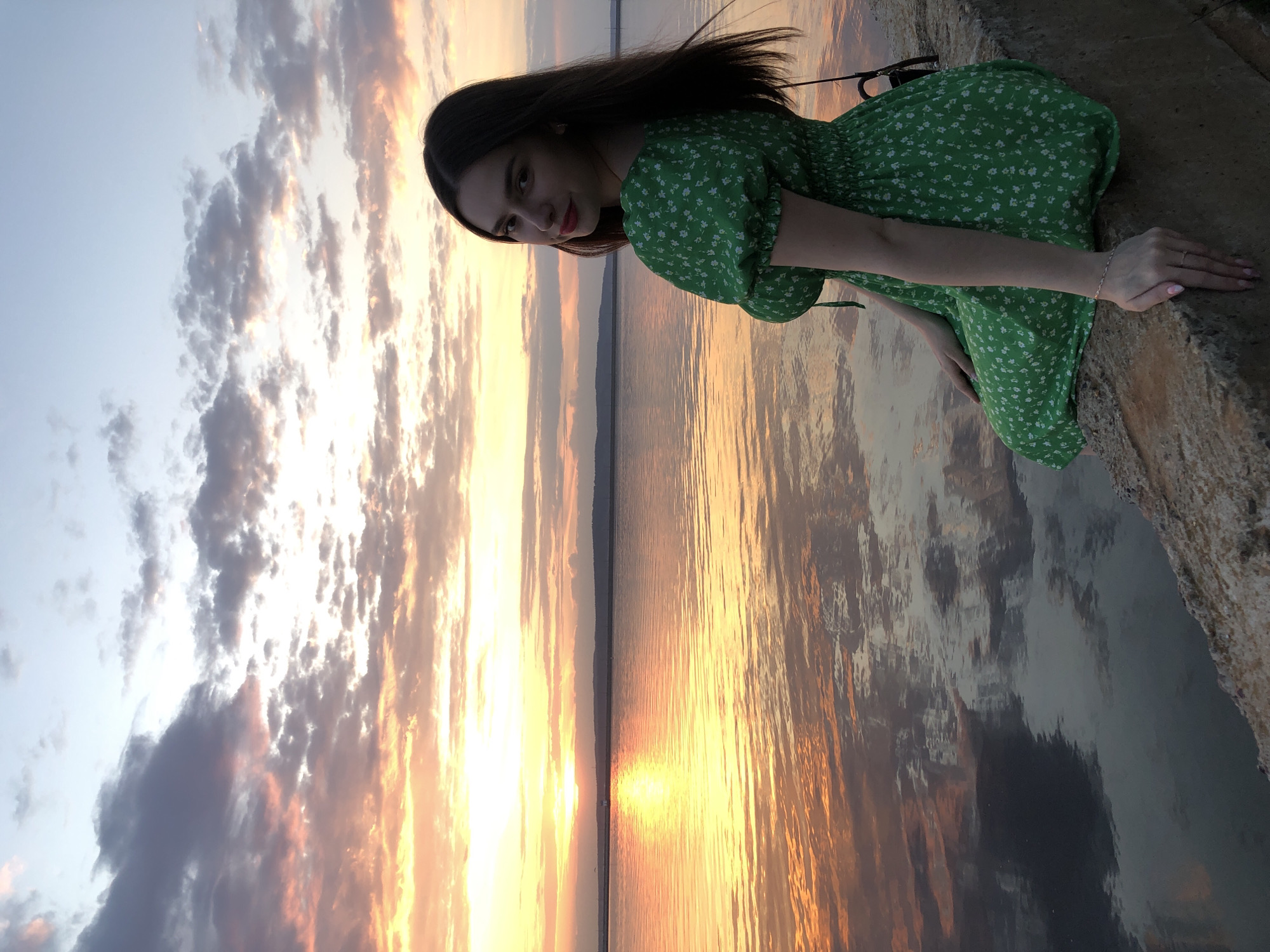A woman in a green floral dress is sitting on a concrete edge overlooking a large body of water at sunset. The sky is filled with orange and grey clouds, reflected in the water. The location is likely in the city of Chaikovsky, Russia.