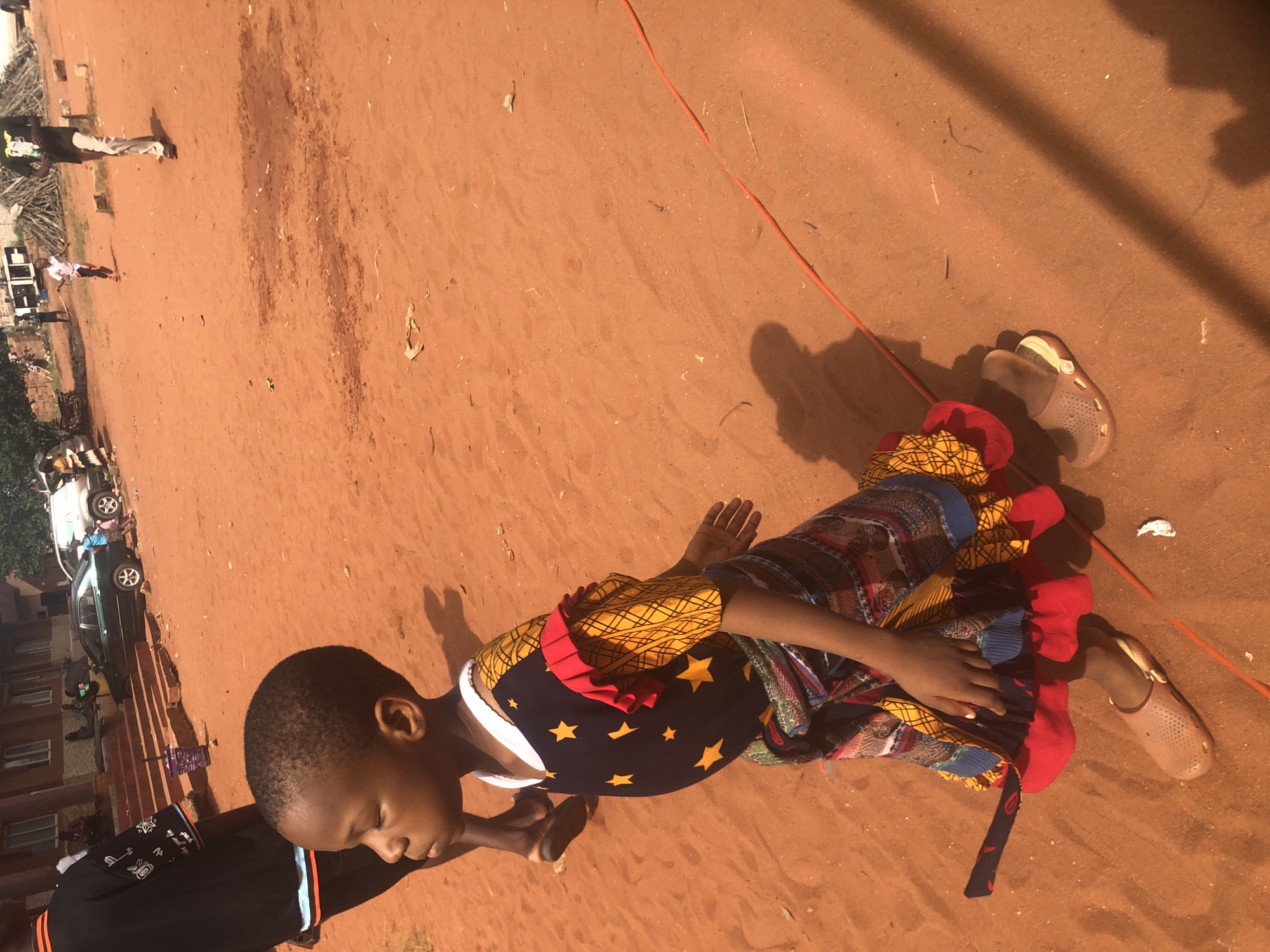 A child lies on their side on the red dirt ground in Nteje, Nigeria. The child is wearing a colorful, patterned dress and tan-colored sandals. An adult arm reaches into the frame from the left, and other people and buildings are visible in the blurred background. An orange cord lays on the ground near the child.