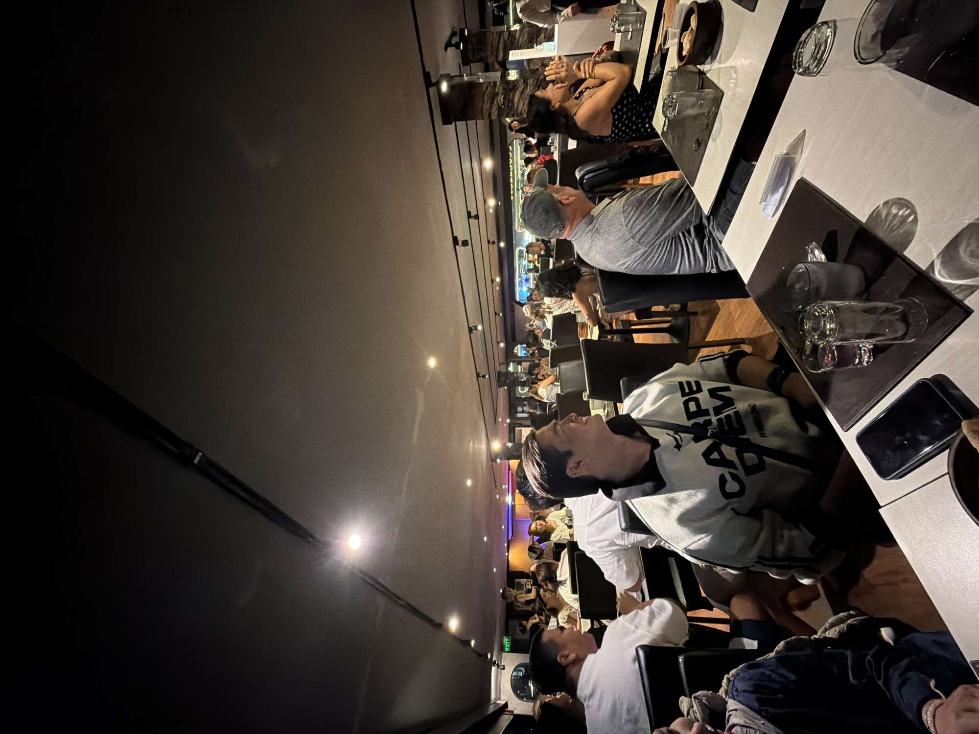 The image captures an indoor scene at a restaurant, likely in Bangkok, Thailand, as suggested by context. The dining area is dimly lit, with a suspended ceiling featuring track lighting and some recessed spotlights illuminating the space. A long, dark structure with a textured surface, possibly a bar or decorative wall, runs along the right side of the room. The scene is populated by numerous patrons seated at tables, engaged in dining and conversation.
In the foreground, several tables are set with white tablecloths, dark placemats, silverware, and glassware. On one table, a smartphone rests on a dark placemat, adjacent to a small bowl containing what appears to be bread.
Several individuals are visible in various states of engagement. A man in the immediate foreground, wearing a light-colored sweatshirt with "CARPE DIEM" printed on it, is seated and looking towards the right. To his left, another person, partially visible and wearing a dark jacket, is also seated. In the middle ground, a man in a grey t-shirt and a grey cap is looking away. To his right, a woman with dark curly hair, wearing a black polka dot dress, is also seated and appears to be interacting with someone or observing something. Further into the restaurant, more diners are seated at tables, with some staff members visible, including a person standing near an "EXIT" sign. The overall atmosphere suggests a social gathering or a mealtime at a moderately busy establishment.