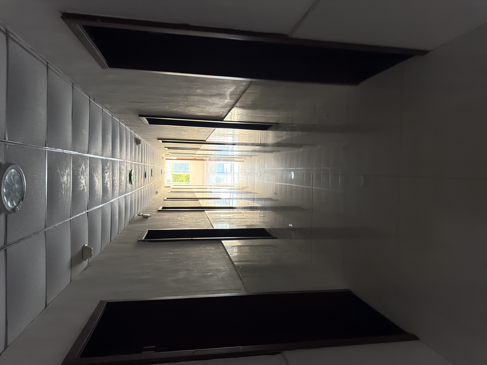 This is a wide-angle, eye-level photograph of an interior hallway with a vanishing point perspective. The hallway is illuminated by overhead fluorescent lights and natural light from a window at the far end. The walls are painted a light, neutral color and appear to be textured. Numerous doors line both sides of the hallway, with dark wooden frames and dark, solid doors. The floor is tiled with a glossy, reflective material that mirrors the ceiling lights and the doors. The ceiling is a grid of acoustic tiles with embedded circular light fixtures. In the foreground, the hallway is darker, with shadows cast by the doors. As the hallway recedes into the distance, it becomes brighter due to the natural light. The image does not depict any people, actions, or interactions. The location is identified as the city of Jiangjin, China.