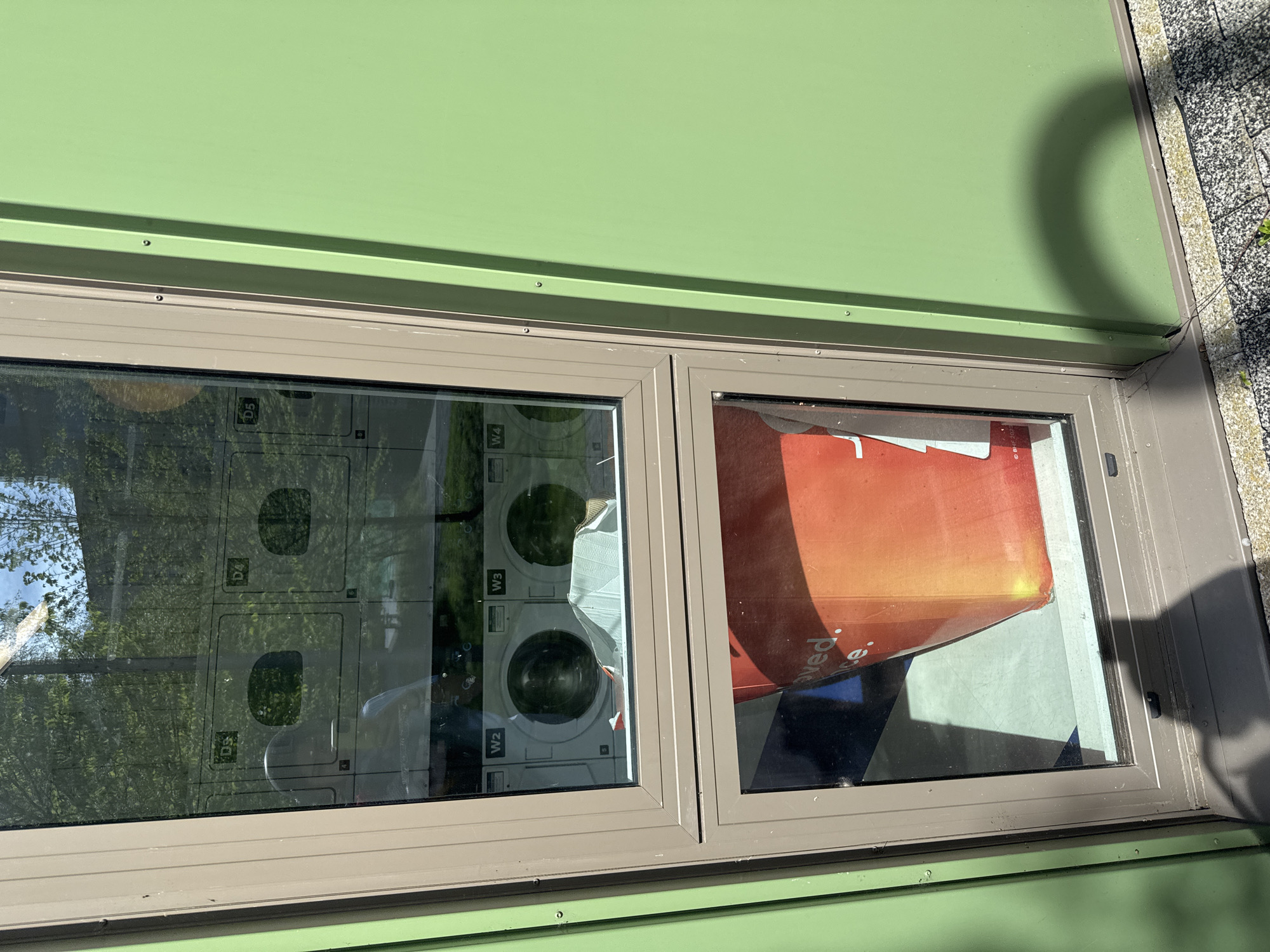 A ground-level, eye-level shot captures a windowed exterior wall, likely of a commercial or service establishment. The wall is painted a light green color with a horizontal dark beige trim supporting two windows. The left window reflects an interior view of a laundromat with multiple washing machines labeled with alphanumeric codes such as "D4," "D5," "W2," "W3," and "W4." The reflections on the window surface are superimposed on the interior view, with tree foliage and light reflecting off the washing machines. The right window reflects an indoor scene with a partially visible red and white promotional advertisement sign. The bottom right corner of the frame reveals a section of light-colored concrete or paving stones. The location is speculated to be in Hull, United Kingdom.