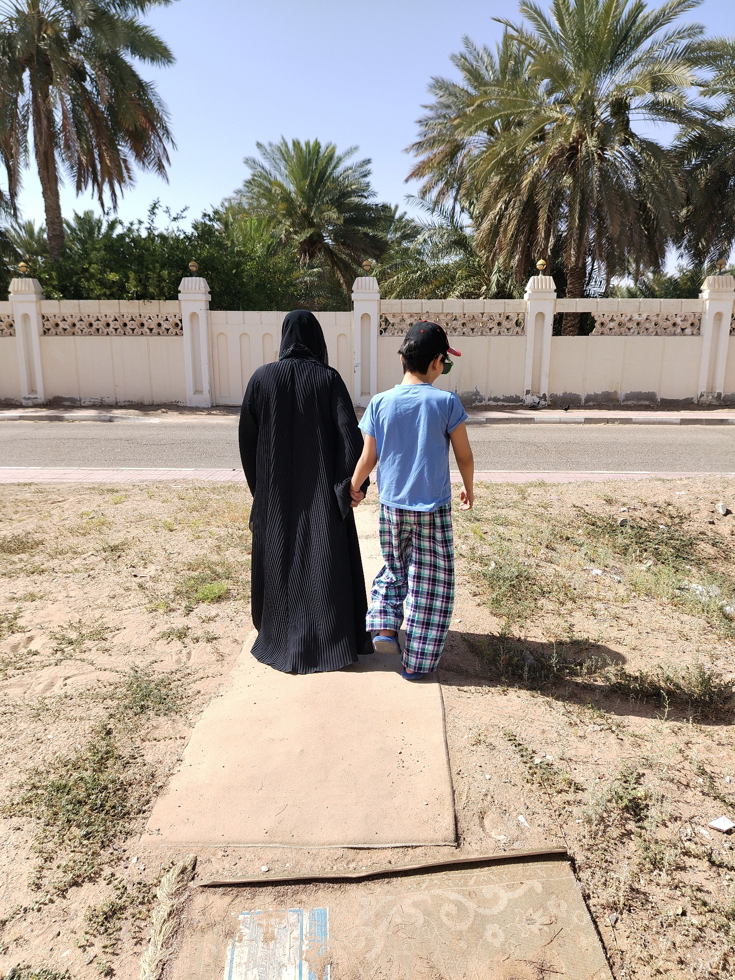 An eye-level, medium outdoor shot depicts a person in a full black abaya holding hands with a child wearing a blue t-shirt, plaid pajama pants, and a red and black baseball cap, viewed from behind. They walk along a narrow concrete sidewalk in the Al Mas'oudi area of Al Ain, United Arab Emirates. The sidewalk is bordered by sandy ground with sparse vegetation. A paved road runs parallel to the sidewalk, with a low, ornate white wall visible beyond. Palm trees line the area behind the wall under a clear, bright sky.