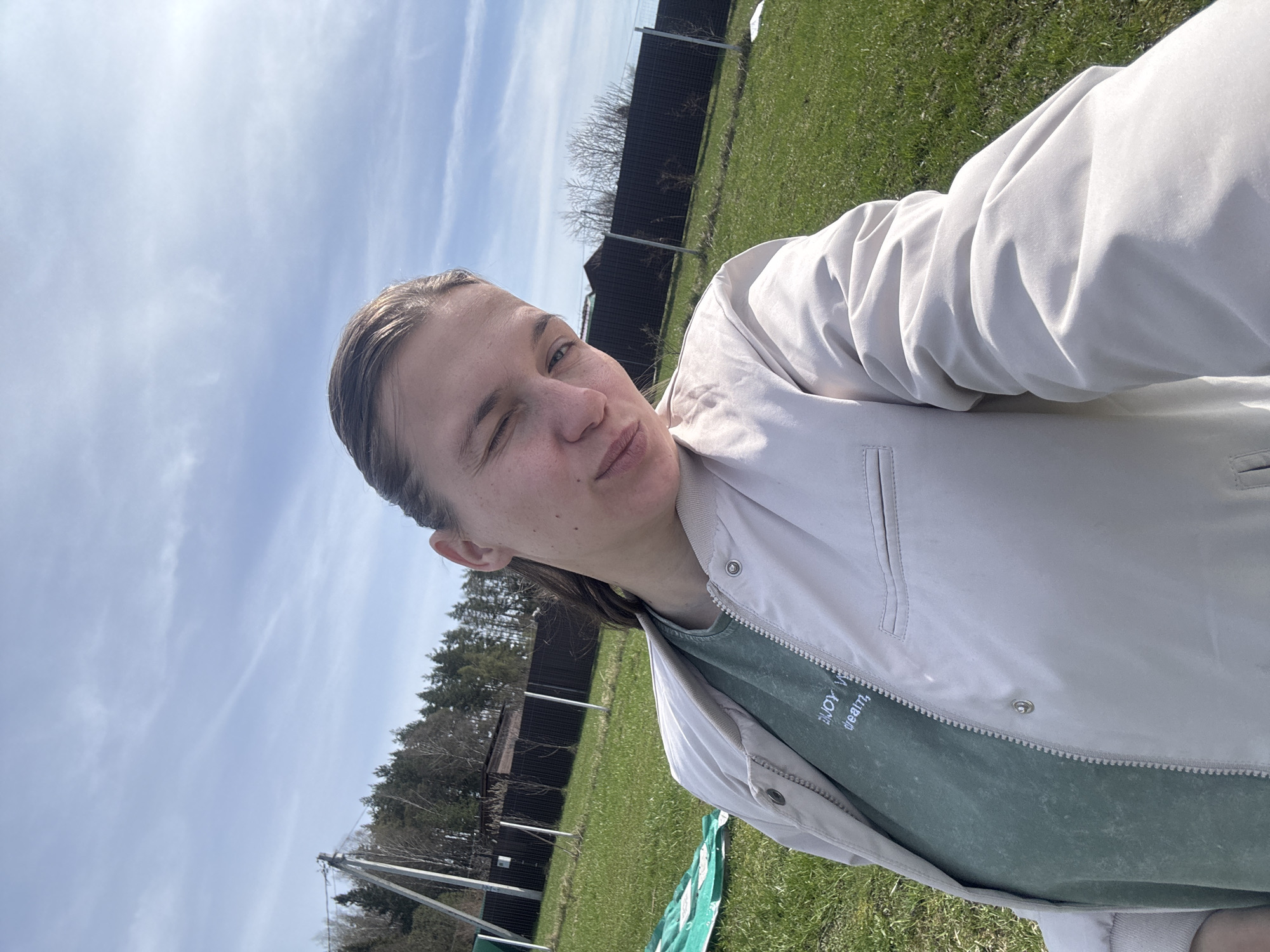 The image captures an outdoor selfie on a bright, sunny day, featuring a person winking with their left eye and a slight pout, suggesting a playful or relaxed expression. The individual has light skin and short or pulled-back dark hair. They are wearing an unzipped light beige or off-white jacket over a dark green t-shirt. White text is visible on the t-shirt, which reads "ENJOY Y" followed by "dream," likely part of a phrase like "ENJOY YOUR dream."
The background shows a clear blue sky with sparse, wispy white clouds. Below the sky, a vibrant green grassy area stretches across the frame. A dark fence, possibly made of metal or dark wood slats, runs horizontally in the middle ground, separating the grassy foreground from a more distant area. Beyond the fence, a mix of bare deciduous trees and dark green evergreen trees are visible, along with a faint building structure on the left side. A light-colored pole, possibly a utility pole, can also be seen in the distance on the left. A crumpled piece of green fabric is lying on the grass in the bottom left corner. The setting appears to be a rural or suburban environment in Razdolye, Russia.