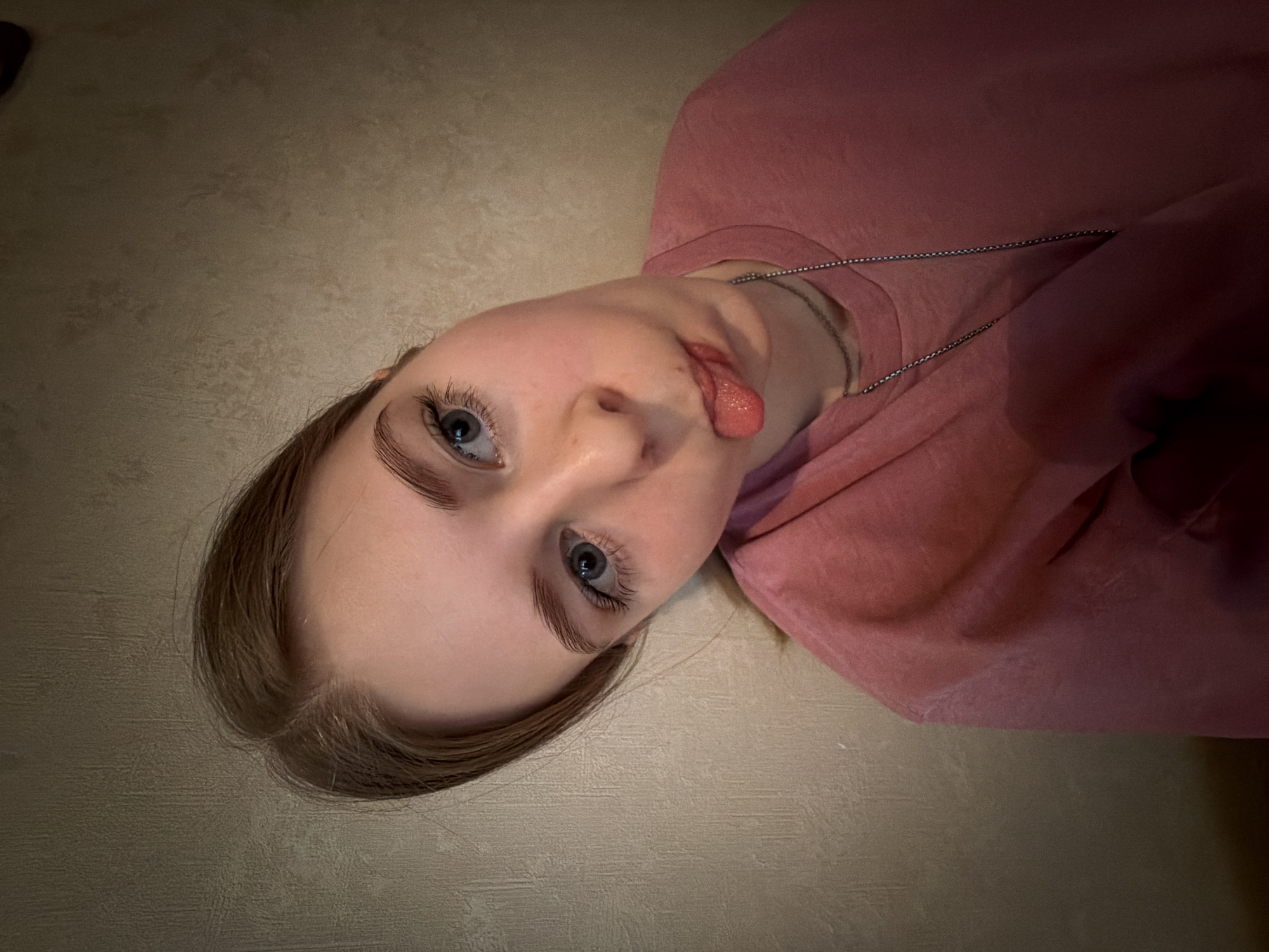 A young fair-skinned person with blue eyes and light brown hair is lying on their back against a neutral-colored surface, possibly a floor. The person is wearing a pink shirt and a silver chain necklace. They are sticking out their tongue. The location is unknown; there is no information in the image suggesting it is Gamovo, Russia.