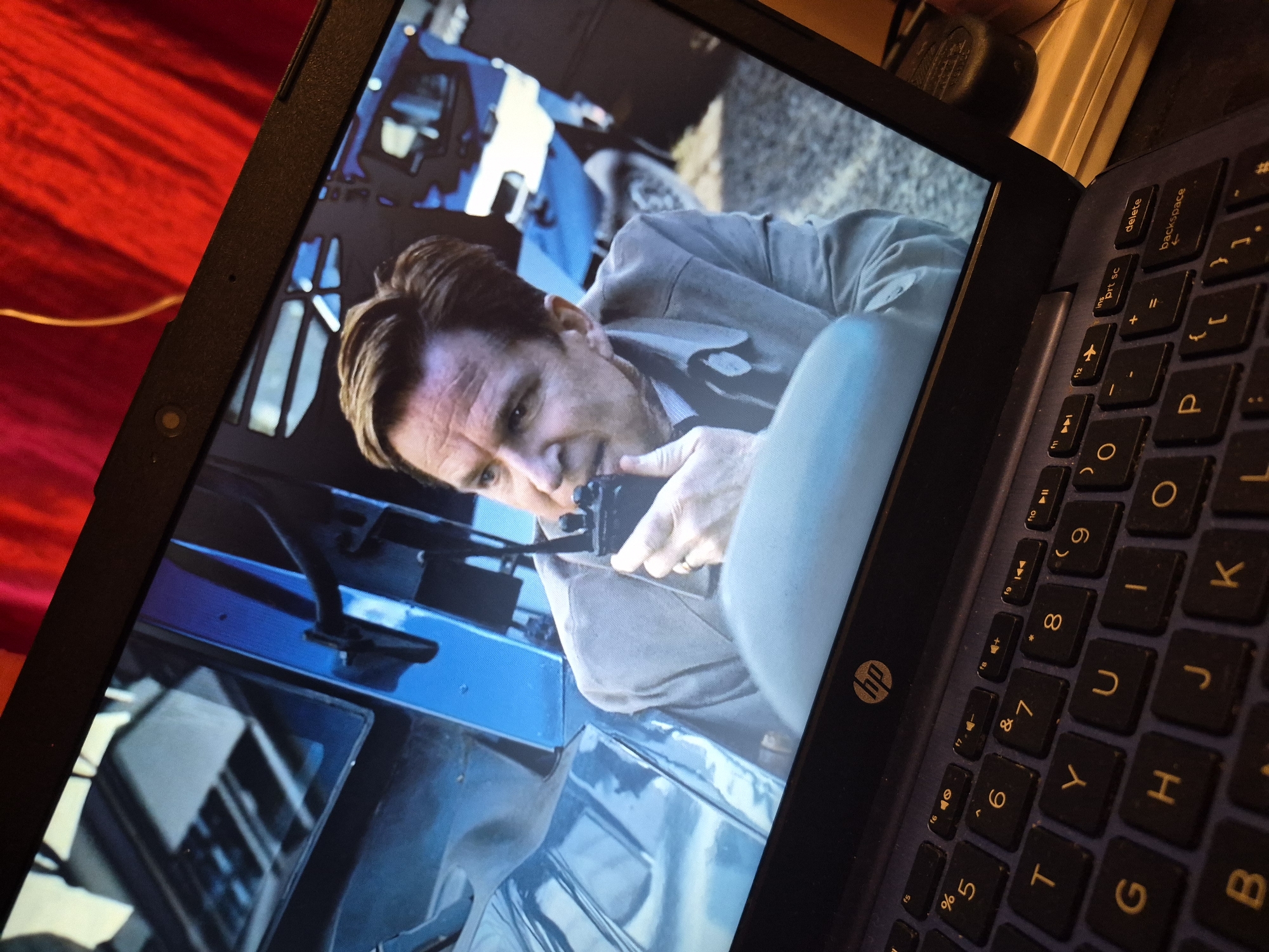 A close-up, angled shot shows a laptop screen displaying a man holding a black walkie-talkie to his mouth. The man, in his late 40s to early 50s, has light skin, brown hair, and is wearing a light-colored button-up shirt. He is seated in what appears to be the interior of a vehicle, indicated by the blue dashboard and steering wheel visible in the background. The overall scene on the screen is dimly lit, with a red curtain partially visible in the top right corner. The laptop itself is a dark color with a visible HP logo. The laptop's keyboard is also in view, with black keys and yellow lettering. The image is taken from a high angle, looking down at the laptop. The exact location in Sandwell, United Kingdom, is not discernible from the image content.