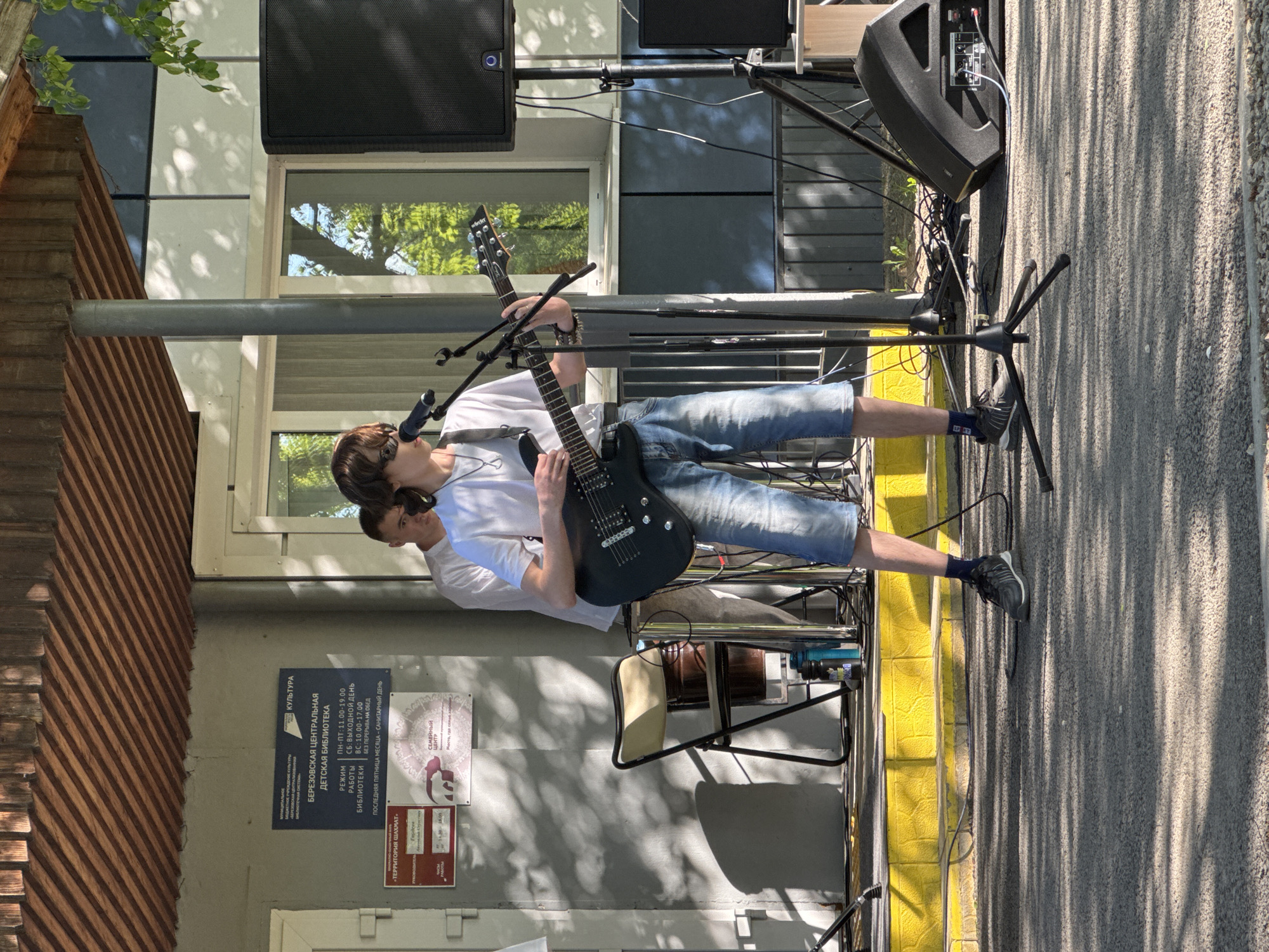 Two young men stand on a sidewalk and play electric guitar and sing into a microphone. They are in front of a building, which has a sign on the wall in Russian for the Berezovskaya Central Children's Library (Березовская Центральная Детская Библиотека), in the city of Berezovka, Russia. A set of speakers are positioned on stands behind them. One of the men is slightly behind the other, but both are wearing white t-shirts and jeans shorts.