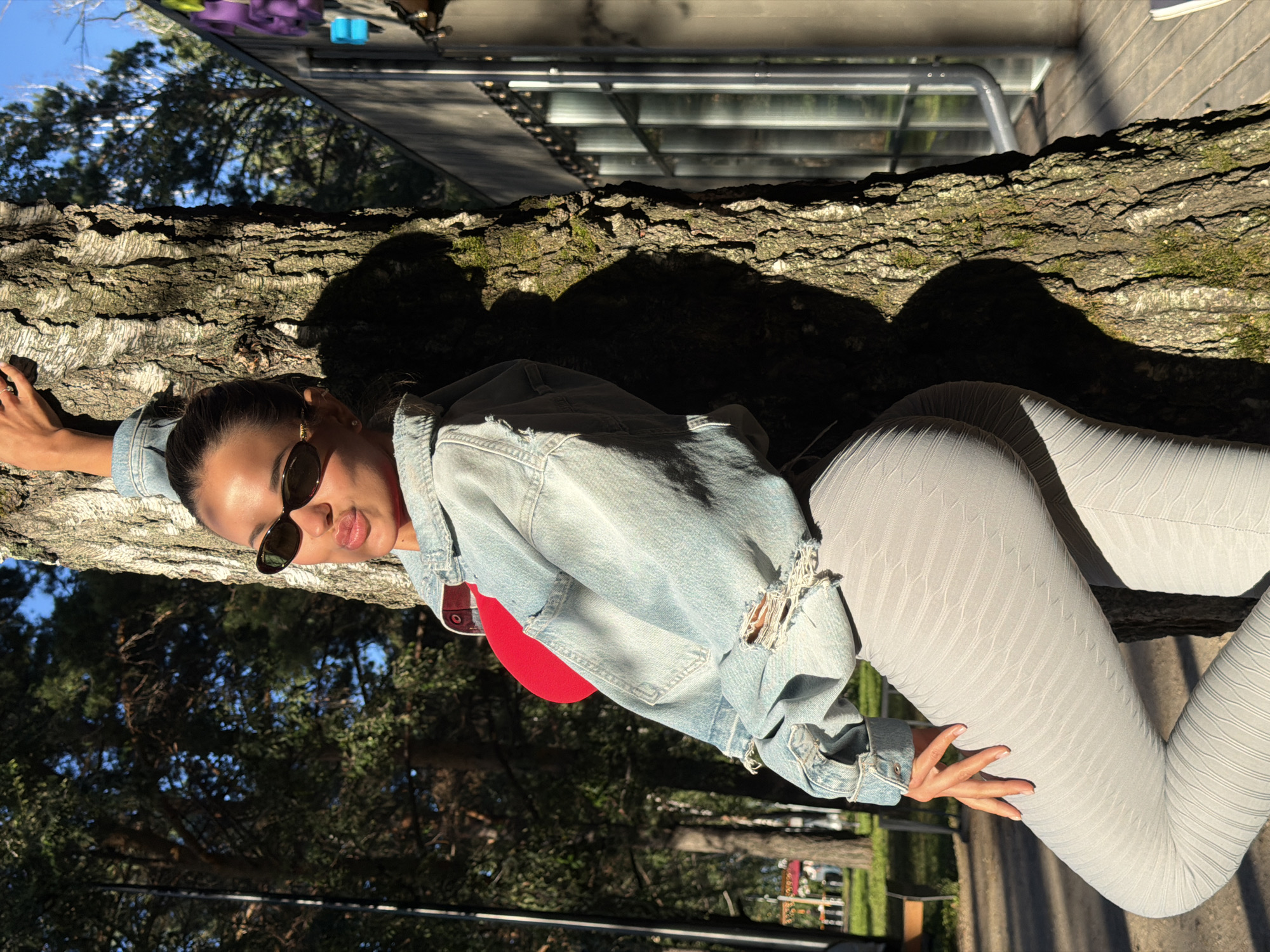 A woman with brown hair pulled back in a bun, wearing sunglasses, a denim jacket, a red top, and gray leggings, leans against a tree. She is outdoors in a park setting, with trees and a building visible in the background. The sun is shining, casting shadows. This image was taken in Krasnoyarsk, Russia.
