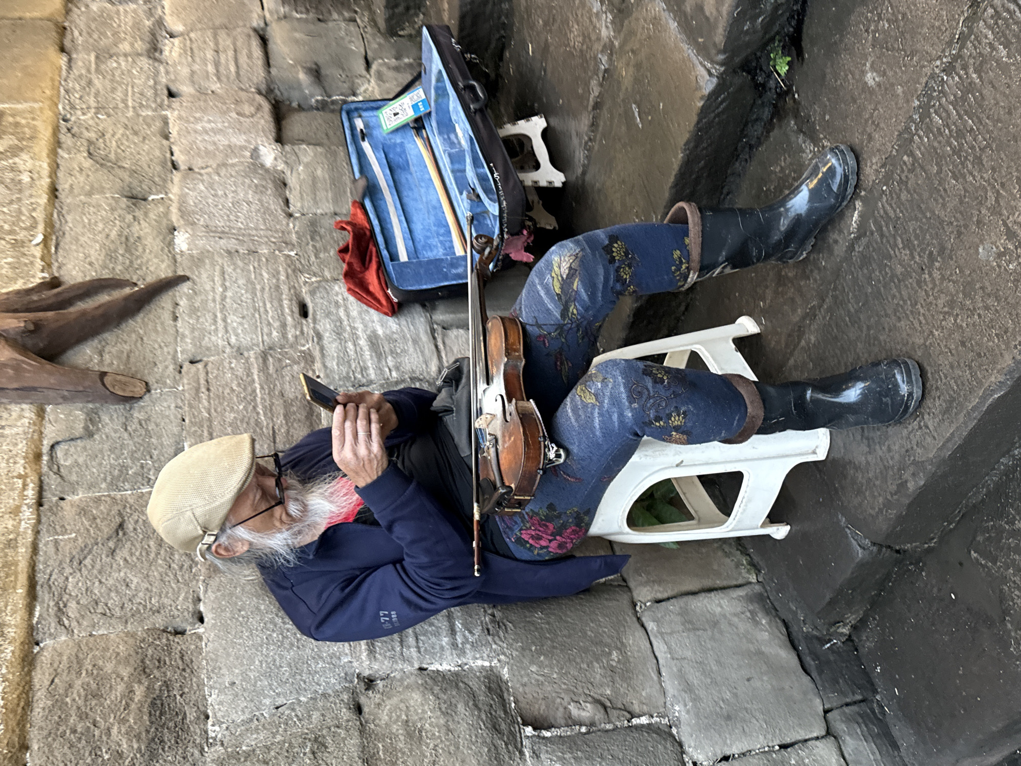 A street musician, likely an elderly man, sits on a small white plastic stool on a stone pavement in Shapingba District, China. He wears a beige newsboy cap, glasses, a long white beard, a dark blue jacket, blue floral-patterned trousers, and black rubber boots. He holds a violin in his hands, poised to play. Beside him is a blue violin case, open to reveal a bow and other accessories. He is positioned against a stone wall on one side and a damp, paved area on the other. A wooden carving is partially visible to his left.