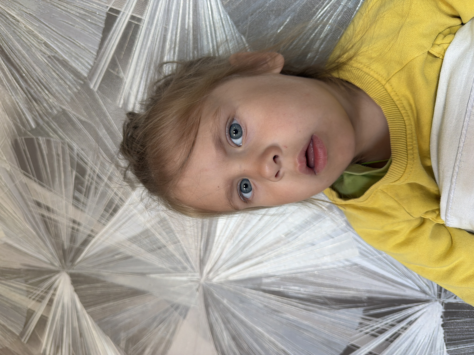 A close-up, eye-level, vertical shot shows a young Caucasian girl with blonde hair pulled up in a messy bun, her bright blue eyes looking directly at the camera with a slightly surprised expression. She is wearing a yellow long-sleeved shirt over a green undershirt, and a white cloth is visible underneath her chin. The background features a textured wallpaper with a radiating, sunburst-like pattern in shades of gray and white. There is no discernible location in the city of Chaikovsky, Russia. The image captures a portrait of the child with a focus on her facial expression and upper body.