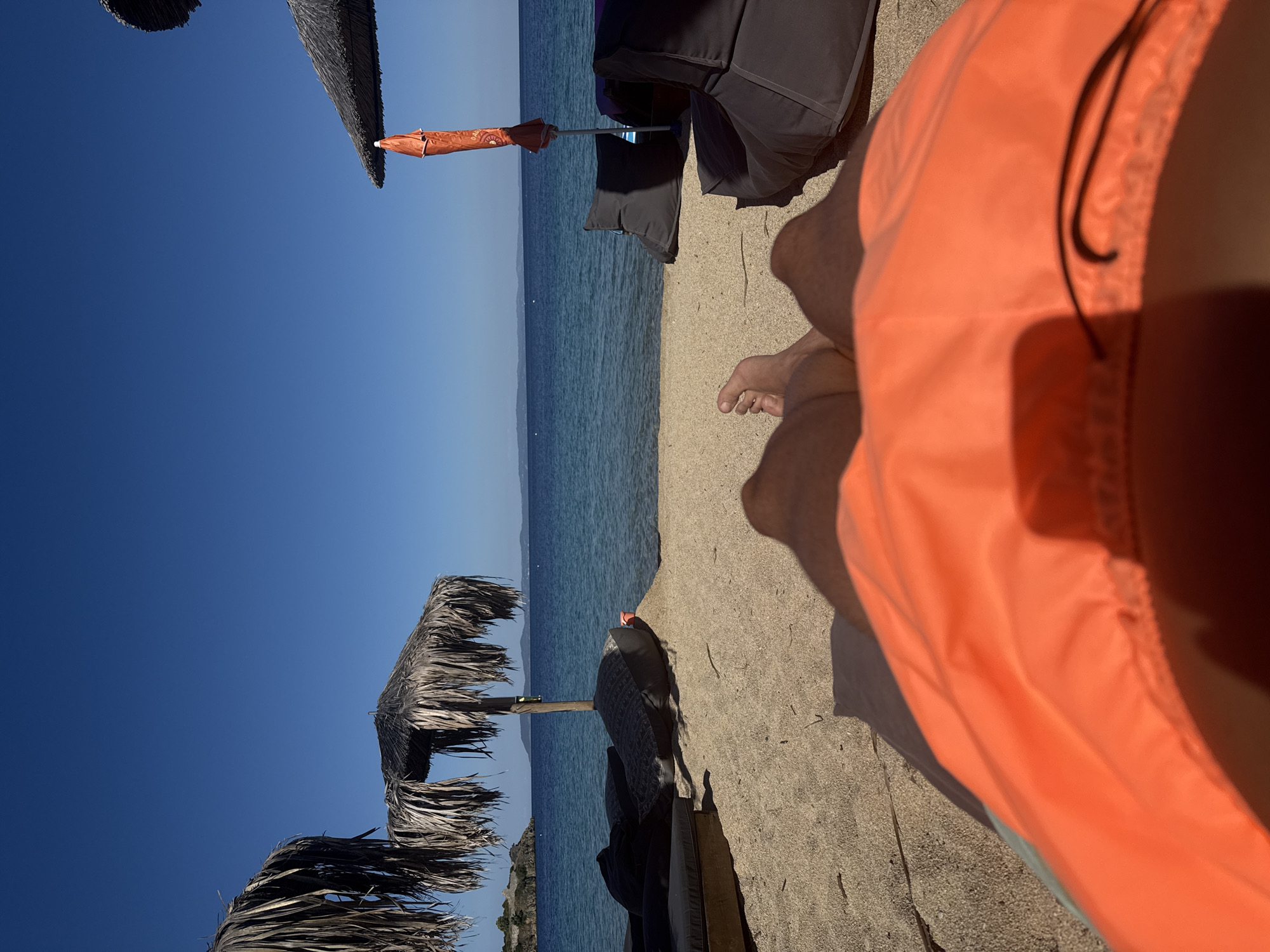 A first-person perspective shot captures a sandy beach in Toroni, Greece, under a clear blue sky. The foreground is dominated by the edge of orange swim trunks. Beyond that, a person's bare legs and feet are visible, resting on the sand. Beach chairs, some covered in dark fabric, and straw umbrellas are scattered across the sand. In the distance, the calm blue sea stretches to the horizon, meeting the sky. The image conveys a relaxed, vacation atmosphere.