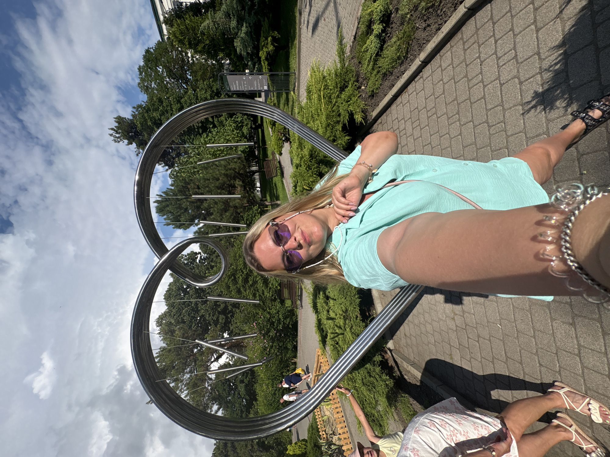 A woman is taking a selfie in front of a large, heart-shaped metal sculpture with hanging chimes in Abrau-Dyurso, Russia. She is wearing a turquoise shirt, sunglasses, and sandals. The sculpture is in a park with trees, bushes, and a paved walkway. In the background, other people are walking and sitting on a bench. The sky is partly cloudy.
