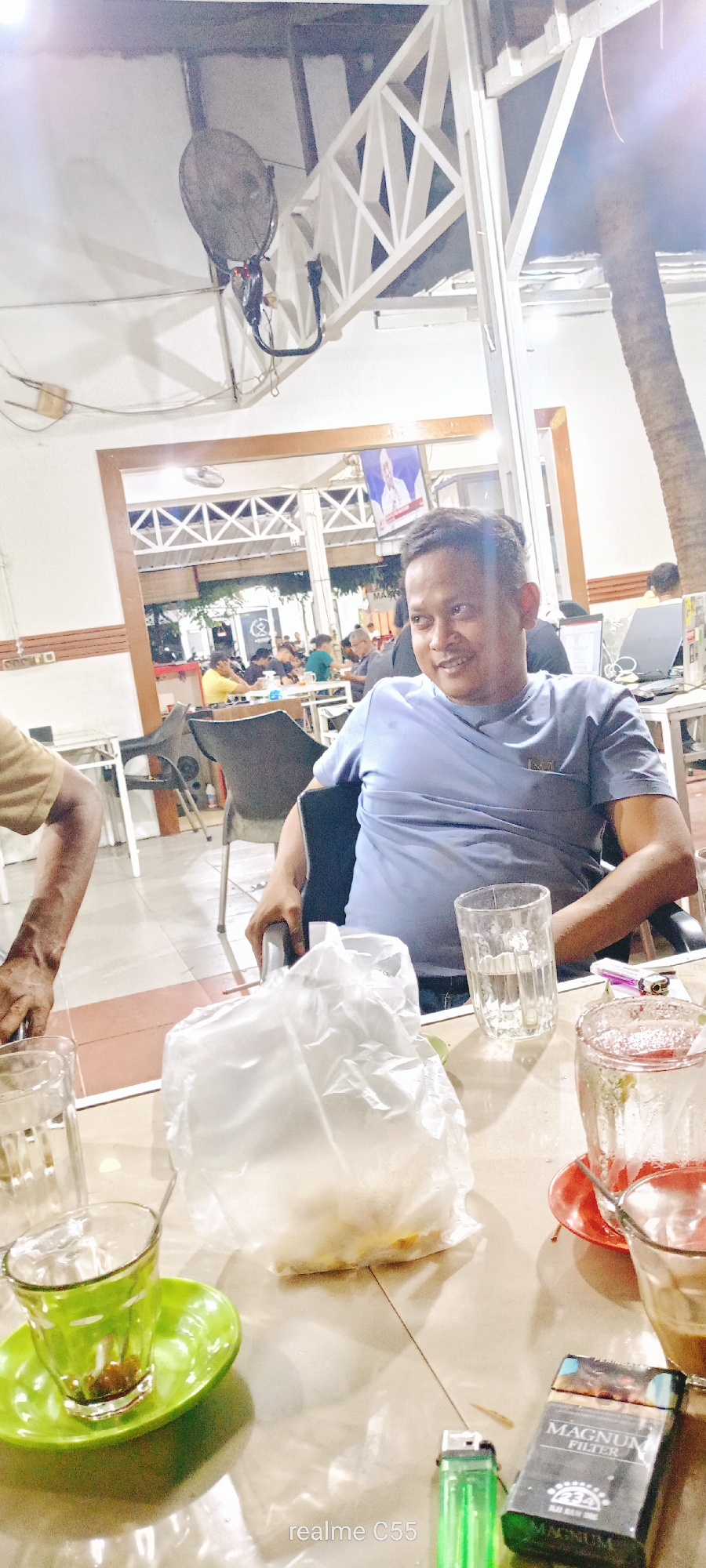 This medium-shot photograph captures a casual evening scene at a cafe or restaurant in Lhokseumawe, Indonesia. The main subject is a man with short dark hair, wearing a light blue t-shirt, seated in a chair. He is smiling and looking towards the right, appearing relaxed and engaged, possibly in conversation. His left arm rests on the chair's armrest.
The table in the foreground holds several items. A white plastic bag, likely containing food such as rice or snacks, is centrally placed. To the right, a clear glass with liquid is visible, along with another glass on a bright green saucer, possibly with tea or coffee residue. A green cigarette lighter and a pack of "MAGNUM FILTER" cigarettes are also on the table. On the far left, a portion of another person's arm, clad in a light brown shirt, is visible.
The background shows a bustling cafe environment with other patrons seated at tables. The setting features white wooden lattice structures and a black ceiling fan, common in open-air or semi-outdoor establishments in warm climates. A large screen in the background displays an image of a person, possibly from a news broadcast or event. A laptop is also visible on a table further back. The presence of artificial lighting indicates the photograph was taken in the evening or at night. A prominent palm tree trunk on the far right further suggests a tropical location. The overall mood is casual and social. A "realme C55" watermark is visible in the bottom left corner.