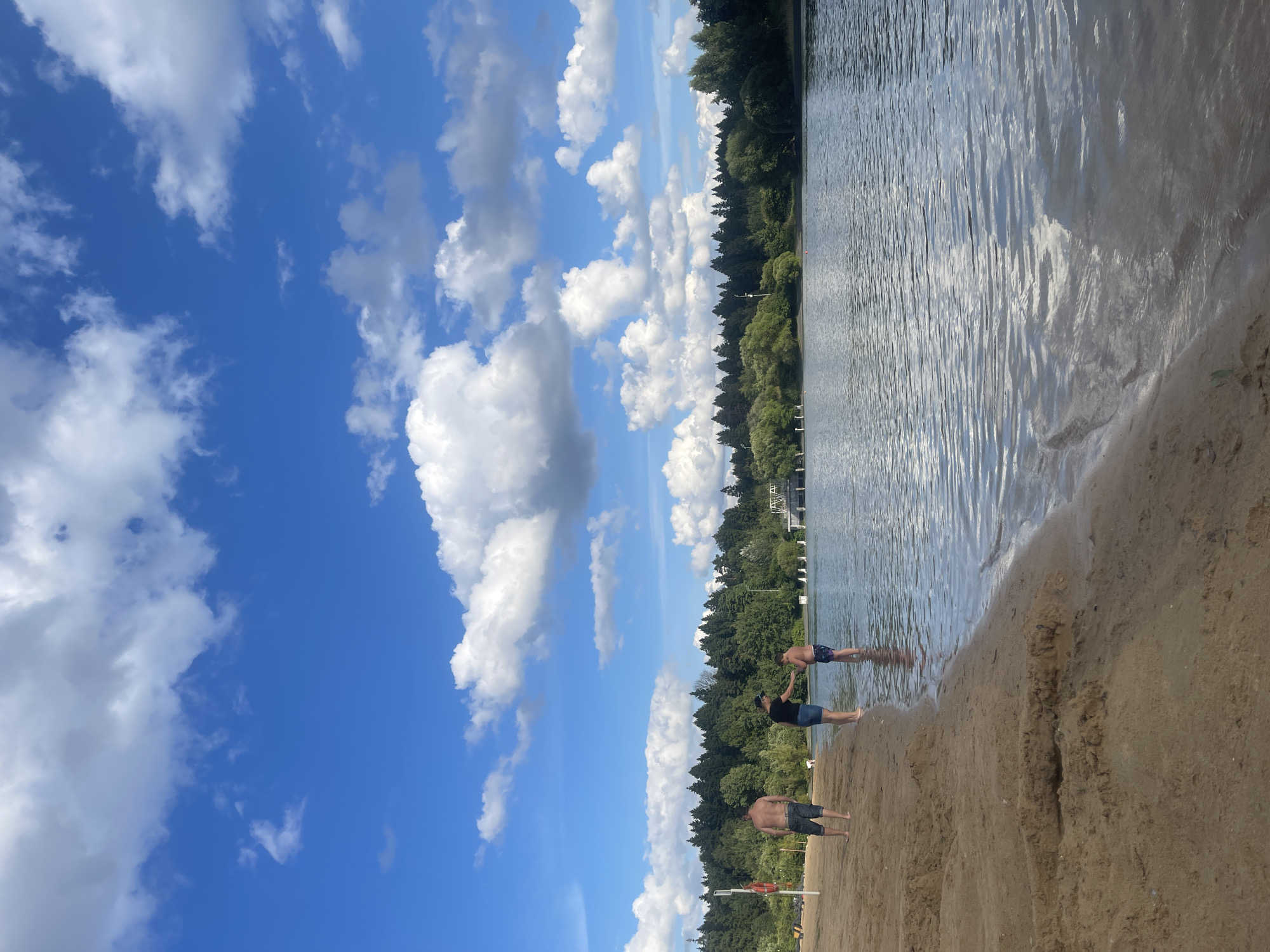 This vertical-oriented image depicts a summer day at a sandy beach by a lake. The sky is a vibrant blue, filled with fluffy white cumulus clouds. The lake's surface is calm, reflecting the sky and a lush green treeline that borders the opposite shore. A few structures, possibly buildings or piers, are visible among the trees.
In the foreground, three individuals are present on the sandy beach. Two men are standing in the shallow water, their legs submerged. A third person, also a man, stands further back on the dry sand. All three appear to be enjoying the warm weather, with casual attire.
The sand is light brown and shows some texture, with footprints and small indentations. The water's edge gently laps onto the shore. The overall scene suggests a leisurely day at the lake, likely during daylight hours, given the bright sky and sunshine. The weather appears to be pleasant and sunny, conducive to outdoor activities. The location is identified as Zelenograd, Russia. There is no visible text in the image.