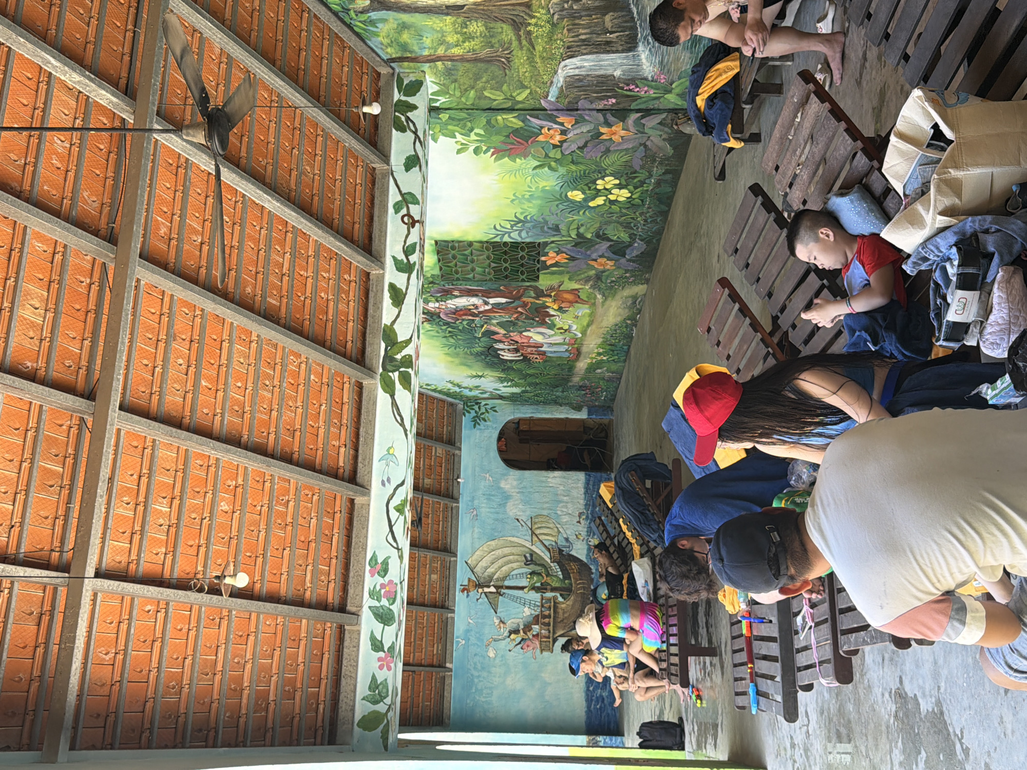 In Tay Nha Trang Ward, Vietnam, an open-air covered seating area features several individuals relaxing on wooden lounge chairs. The ceiling is constructed with orange tiles and exposed beams, with a ceiling fan visible. The walls are adorned with colorful murals depicting a pirate ship in a blue seascape on one side, and a lush forest scene with a waterfall and diverse flora on the other. A few people are seated or reclining, some are looking at their phones, and one child is lying down with a phone. Bags and towels are scattered around the lounge chairs. The floor appears to be wet or reflective concrete. The overall scene suggests a recreational or resort setting.