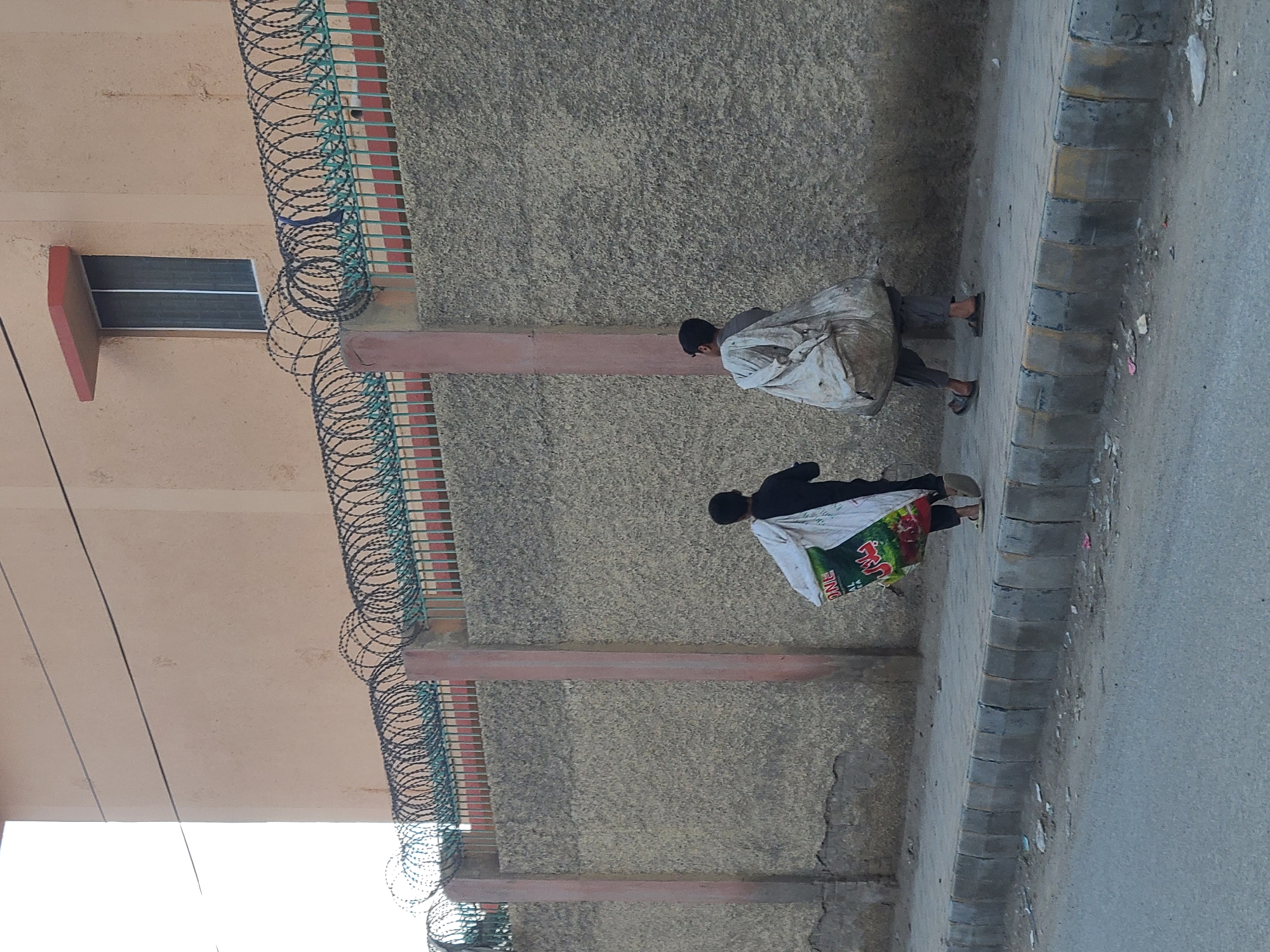 Two individuals walk along a sidewalk next to a building in Quetta City Tehsil, Pakistan. The building has a window and a barbed wire fence at the top. One individual carries a sack over their shoulder, while the other carries a bag with visible text in what appears to be a non-English script. The environment is urban, with a sidewalk and street visible.
