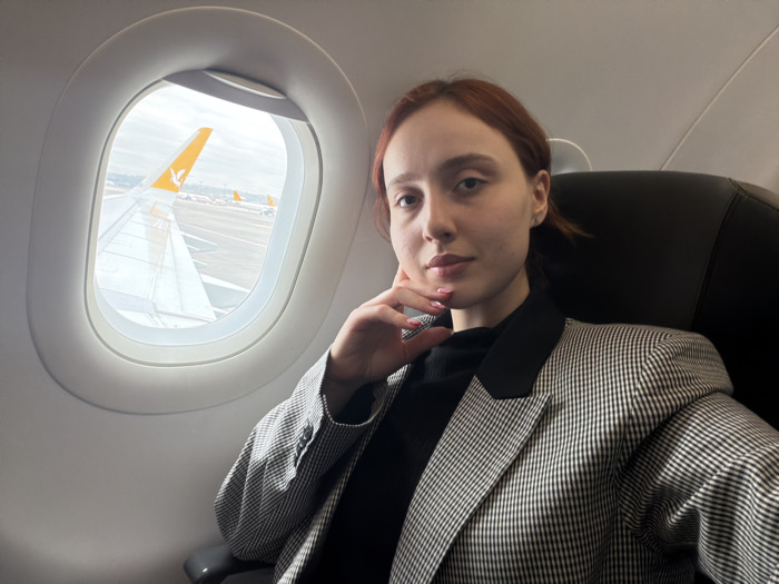 A female individual is seated in an airplane cabin, specifically identified as a Pegasus Airlines aircraft, positioned on an airfield at Sabiha Gökçen International Airport (SAW) adjacent to Tuzla, Turkey. The subject, with reddish-brown hair, is oriented towards the camera. Her right hand, adorned with pink nail polish, rests gently beneath her chin. She wears a black turtleneck garment overlaid with a black and white checkered blazer featuring solid black lapels. To her left, an oval aircraft window displays a white airplane wing in the immediate foreground, obscuring the engine. Beyond the wing, a yellow vertical stabilizer with the Pegasus Airlines logo is distinctly visible on the tarmac, alongside other aircraft and a cloudy sky. The ground visible through the window appears to have patches of snow or frost.