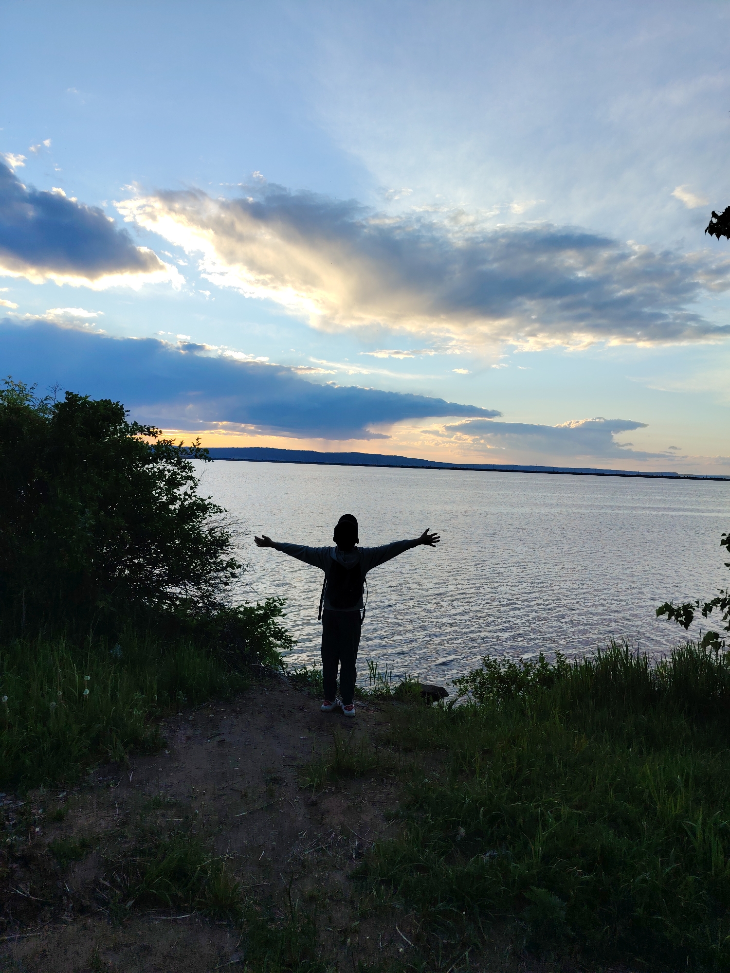 A person stands at the edge of a grassy bank overlooking a large body of water. The person is silhouetted against the evening sky, arms outstretched. The person is wearing dark clothing, including a hooded jacket, and a backpack. The sky is a mix of blue, white, and gray clouds, with the setting sun casting a golden glow. On the left side of the image, there is a patch of bushes and trees. The location is likely in the city of Chaikovsky, Russia, given that it was specified in the prompt. The water appears calm, and there is a distant shoreline visible. The foreground consists of a dirt path and tall grass.