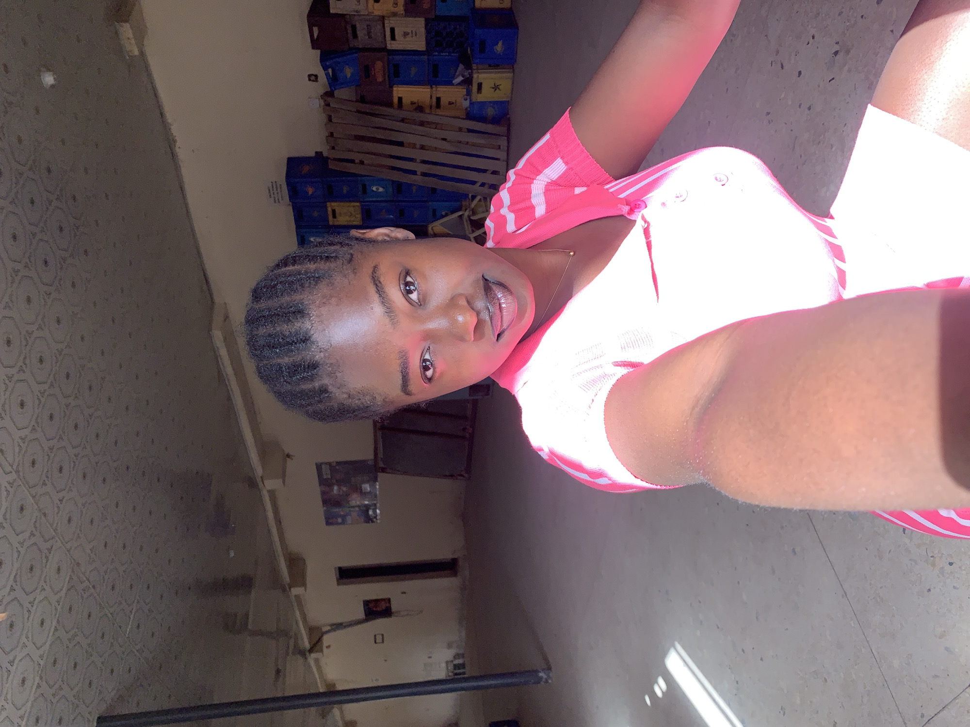 A person is seen indoors, seemingly taking a selfie. The individual has dark hair styled in braids and is wearing a pink and white striped shirt. They are looking at the camera with their mouth slightly open. Behind them, there are stacked blue and yellow crates, possibly stacked soda crates, along a wall. The background also reveals a patterned floor and a wall with what appears to be a poster or sign. Given the prompt's request to include Kabong, Nigeria, it is presumed that the location is a building within the city limits of Kabong, Nigeria. The environment is somewhat dimly lit and the orientation is in portrait.