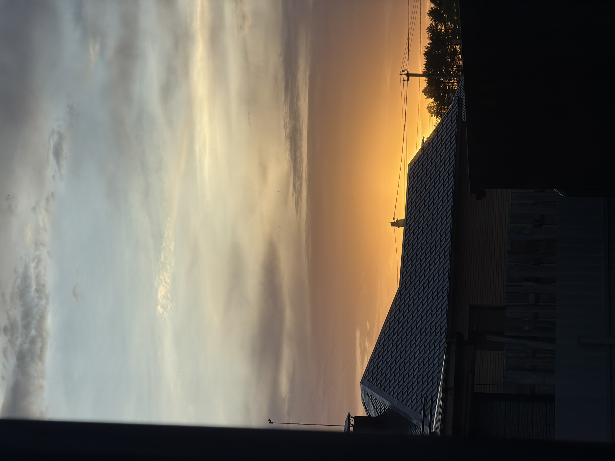 This image depicts a sunset in Chaikovsky, Russia, with a view from what appears to be a window. The sky is filled with clouds that transition from light gray and white on the left to shades of orange and yellow near the center. A silhouette of trees and a power pole with wires are visible against the bright sky. To the right is the dark silhouette of a building with a roof covered in what appears to be diamond patterned roofing materials. The building is partially obscured by darkness. The bottom of the frame is a dark foreground, possibly part of the window or balcony structure from where the image was captured.