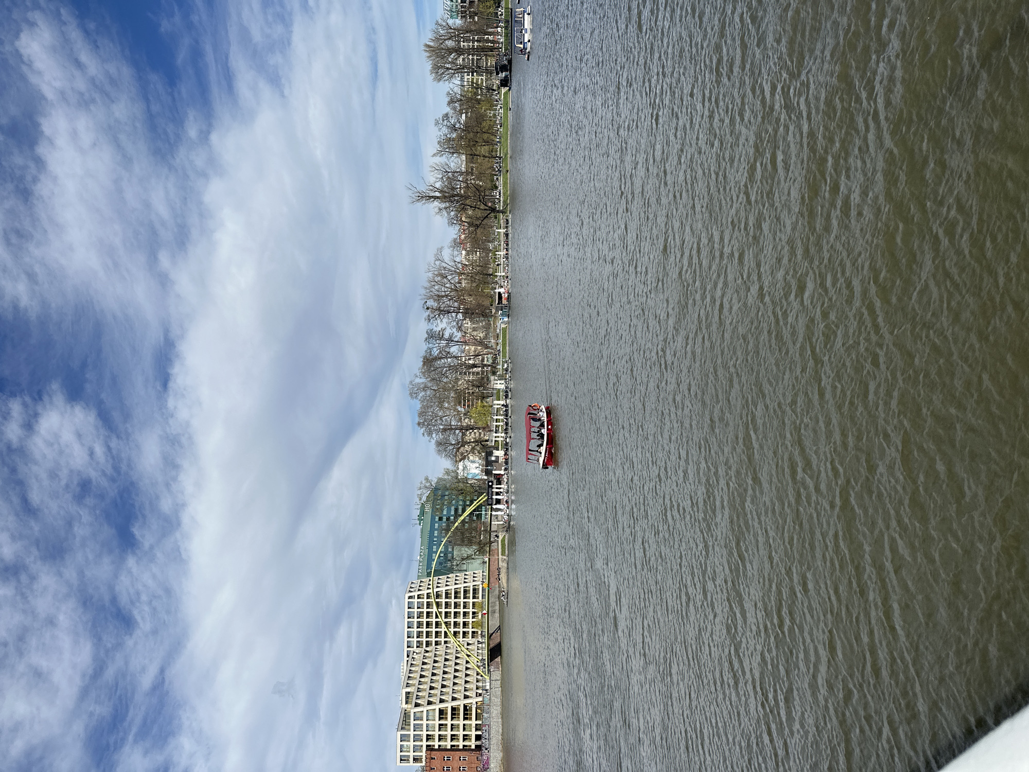The image depicts a body of water, likely the Oder River, flowing through Wrocław, Poland. A red tourist boat with passengers is visible on the river. On the riverbank, there is modern architecture including the Park Plaza hotel and a building with a distinctive grid-like facade. A yellow art installation or bridge-like structure is also seen. Trees line the riverbank, and buildings are visible in the distance. The sky is blue with scattered clouds.