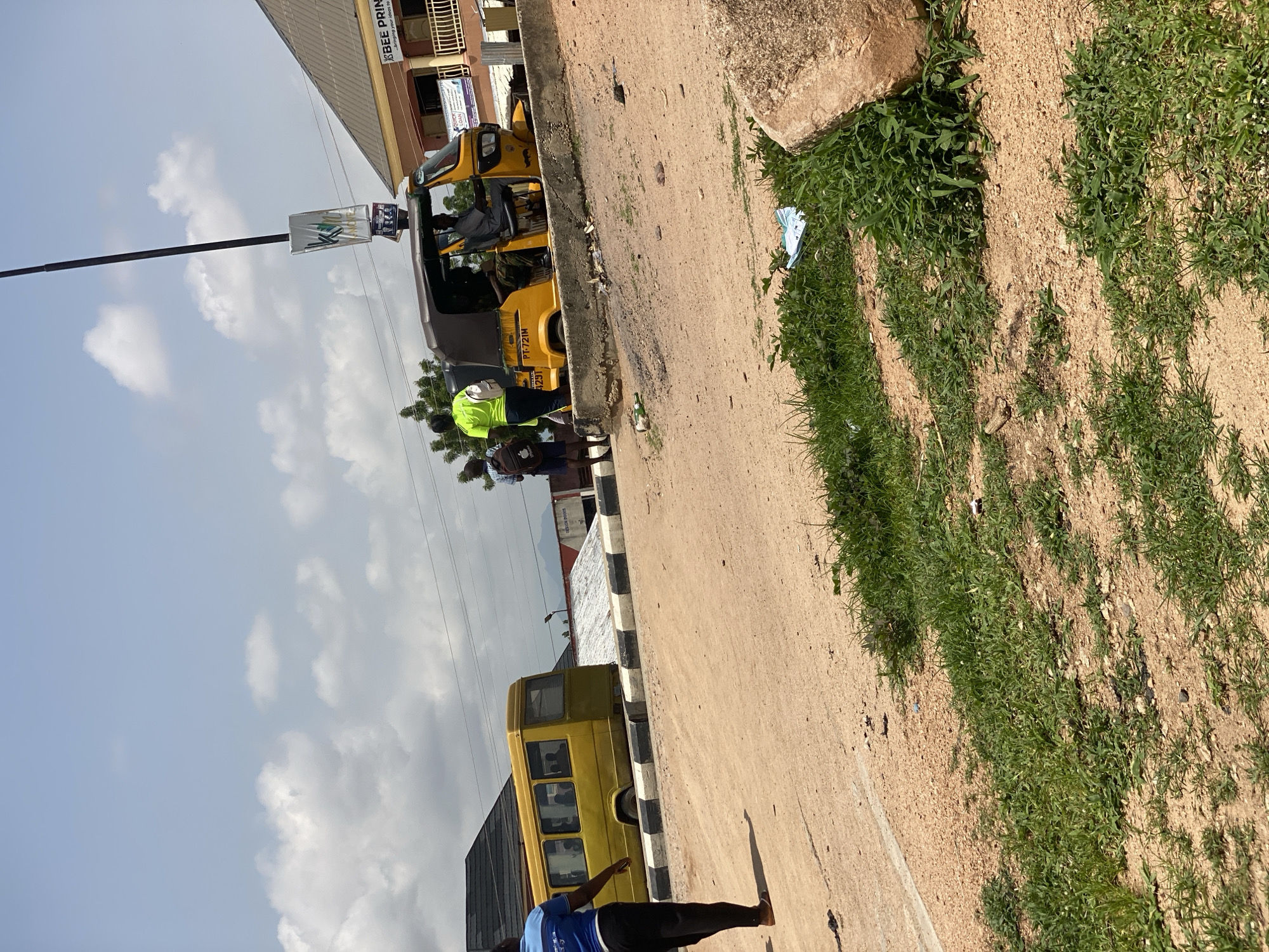 This is a street-level outdoor photograph taken in Jalingo, Nigeria. The scene depicts a dusty, unpaved road with sparse green vegetation and scattered debris. In the foreground, a person in a blue shirt and black pants walks away from the camera towards the left. To the right, a large rock and more green growth are visible.
In the middle ground, a yellow minibus is parked. A short concrete barrier with black and white stripes runs along the side of the road in front of the bus. To the right of the bus, a yellow auto-rickshaw (a three-wheeled vehicle) is stopped, with a driver and at least one passenger visible. Two individuals stand near the auto-rickshaw; one is wearing a bright green shirt and has a backpack, and the other is wearing a blue and white checkered shirt.
In the background, several buildings are visible, including one with a sign that reads "BEE PRINT." A tall pole with a banner hangs in the air. The sky is partly cloudy. The overall impression is of a typical roadside scene in a bustling urban environment.