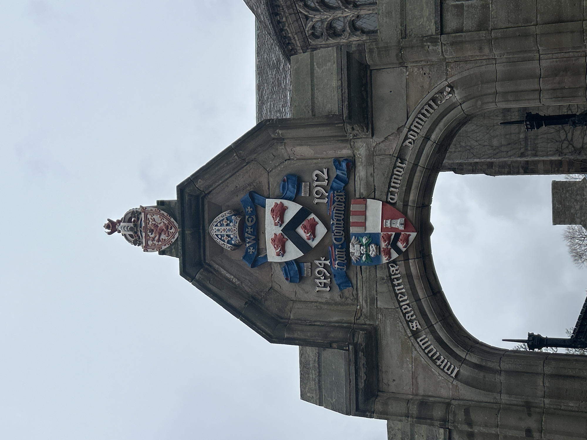 Image depicts a stone archway with elaborate heraldry above it. The archway features Latin inscriptions; "Amor Domini Initium Sapientiae". Above the arch is a heraldic display with multiple shields, including one with three boars' heads, and dates 1494 and 1912. The archway is part of a stone structure, likely a building. The sky in the background is overcast. Location: Likely within the University of Aberdeen, Scotland, United Kingdom.