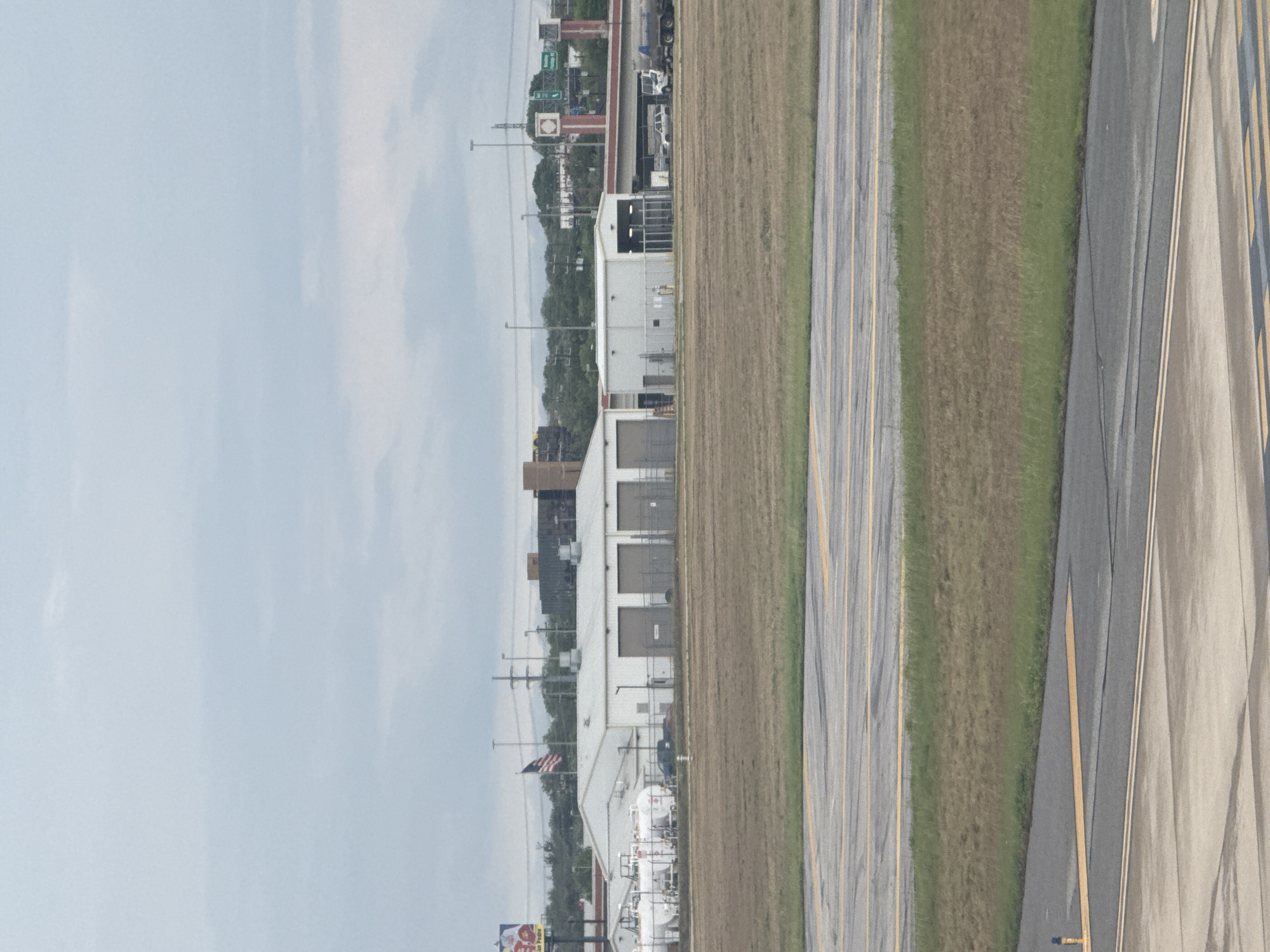 This image depicts a landscape in what appears to be San Antonio, Texas, United States of America. There is an airport with an American flag on top of the airport. The landscape includes airport runways and green space. There are buildings and industrial structures in the background, along with power lines and light poles. A highway and adjacent city street are running in the background. The sky is overcast.