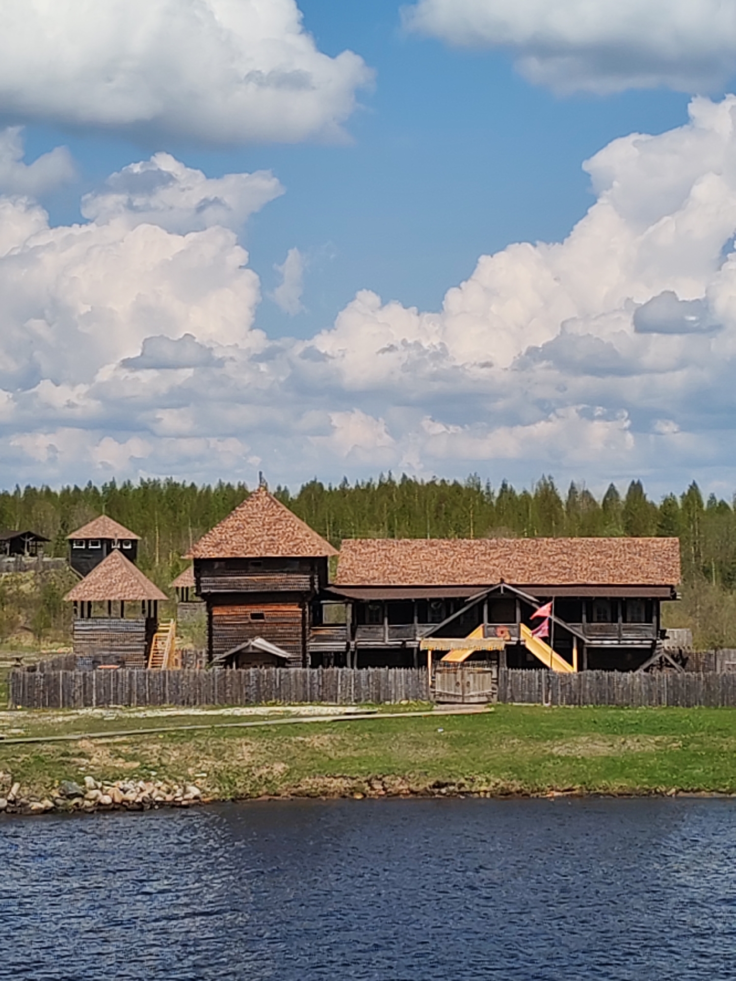 A medium shot depicts a wooden fort complex with multiple structures behind a wooden fence, near a body of water. The buildings have reddish-brown roofs and dark wooden walls, with some having elevated walkways and stairs. The sky is blue with puffy white clouds, and a green forest is visible in the background. A grassy bank is in the foreground, separating the fort from the water.