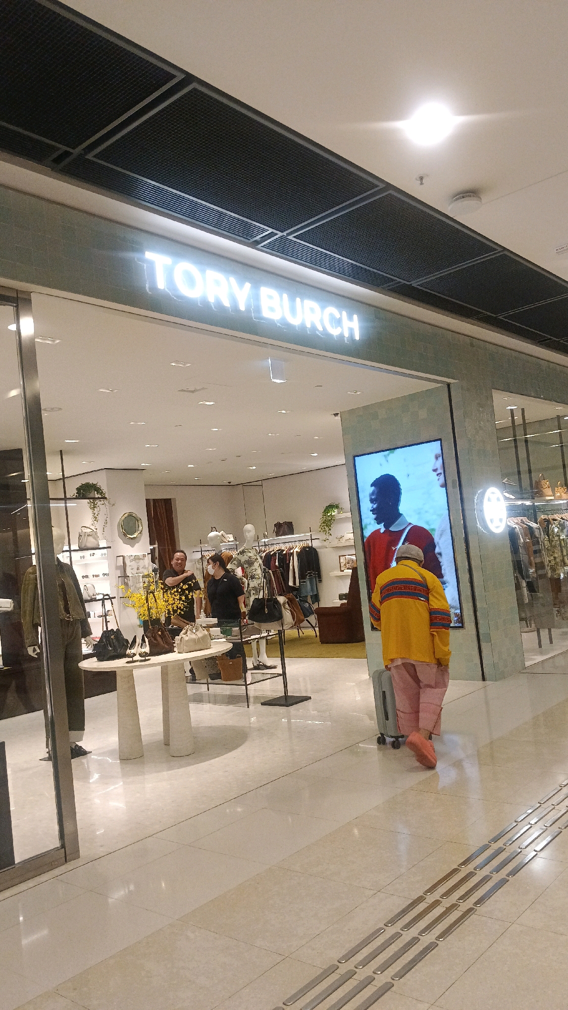 The image depicts the interior of a Tory Burch store, identified as being located in Hong Kong, China, by its prominent signage. The store's facade features a large, illuminated "TORY BURCH" logo. Inside, the retail space is brightly lit and organized, with mannequins displaying clothing and handbags on display tables and racks. Several individuals are present: two staff members are visible behind a counter, one interacting with a customer. A person wearing a bright yellow jacket and pink pants is seen walking away from the camera, pulling a light-colored suitcase, and looking at a large digital screen displaying an advertisement featuring two individuals. The floor is tiled with a light-colored material, and a tactile paving strip is visible in the foreground.