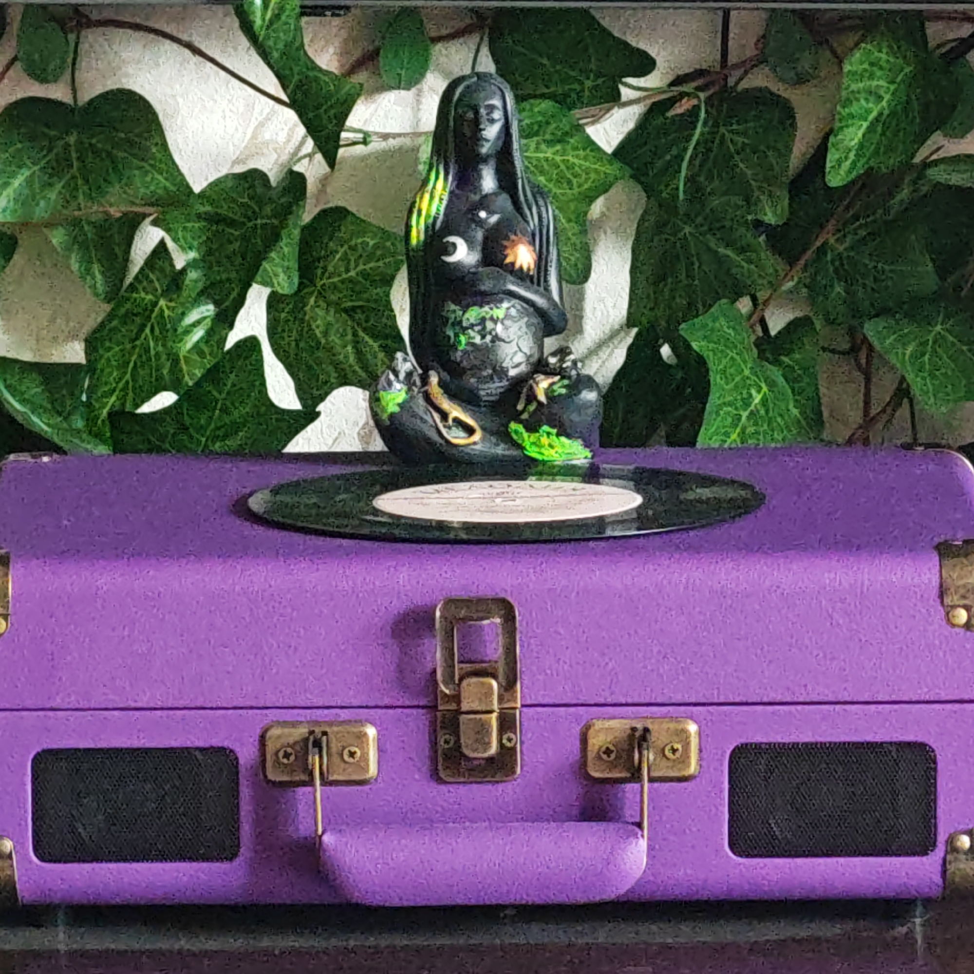 A medium, eye-level shot depicts a purple record player topped with a black vinyl record and a statue. The statue, centrally positioned, appears to be a dark-toned figure with celestial motifs—a crescent moon and sun—on the torso, and green accents at its base and in its hair. The statue sits on the vinyl record which lies flat on the record player. The purple record player, which occupies the lower half of the frame, is detailed with antique gold latches and a handle. Behind the record player and statue, artificial green ivy cascades down from the top of the frame, providing a natural backdrop. The wall behind the ivy is a textured white. The scene is set indoors, with the composition suggesting an artistic or decorative arrangement. The image was captured in Rostov, Russia.