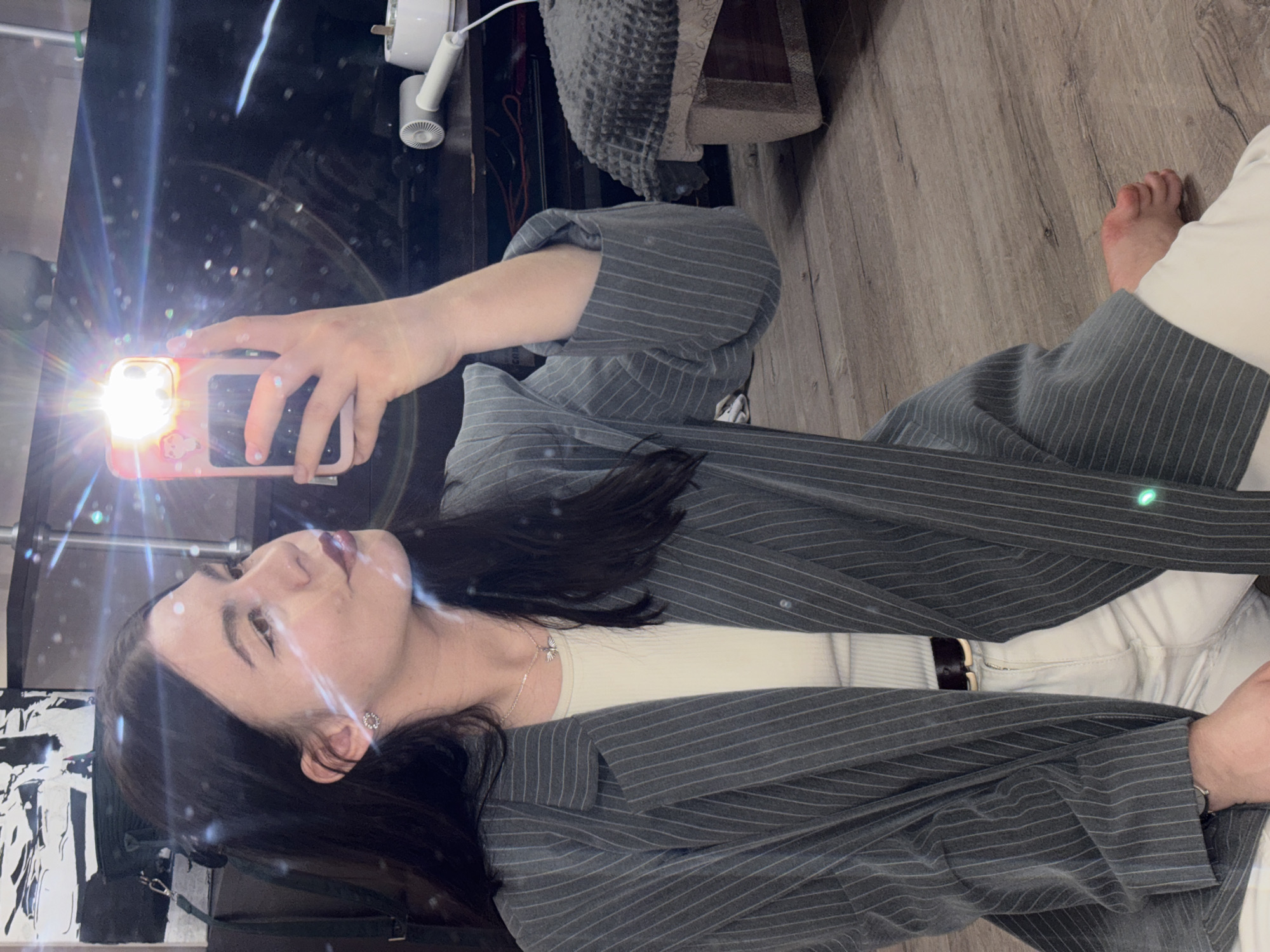 A woman with dark hair takes a selfie in a mirror. She's wearing a gray pinstripe blazer, a white top, and white pants with a black belt. The flash from her phone illuminates the scene. Part of a foot is visible in the background, along with a wooden floor and a gray upholstered item. The environment includes a black surface with reflective elements. There is no evidence suggesting the image was taken in Perm, Russia.