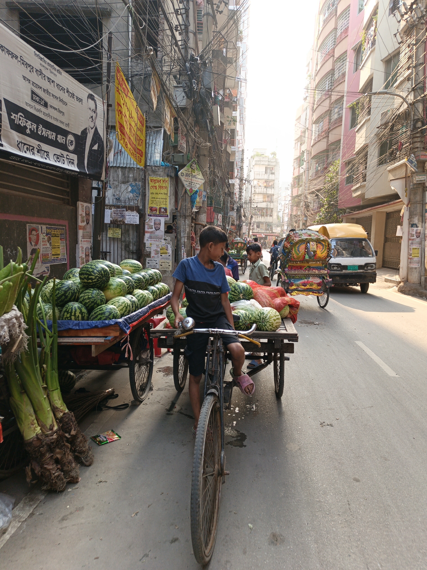 The image captures a bustling street scene in Dhaka, Bangladesh. A young boy is at the forefront, seated on a bicycle attached to a wooden cart piled high with watermelons, ready for sale. Another boy stands beside the cart, seemingly involved in the same trade. Adjacent to the watermelon cart is another smaller cart loaded with watermelons covered with red net. To the left of the watermelon cart are tall green stems of some type of crop.
A crowded urban setting surrounds them, with buildings lining both sides of the street. Overhead, a dense web of electrical wires creates a tangled canopy. Posters and banners adorn the buildings, featuring text and political figures.
Further down the street, a van and an elaborately decorated rickshaw-like vehicle move along. The street itself is paved with a visible painted white lane marking. The overall impression is one of a vibrant, crowded, and busy urban commercial area in Dhaka. The image suggests the location to be around Kafrul-Mirpur as indicated by the election poster on the left.