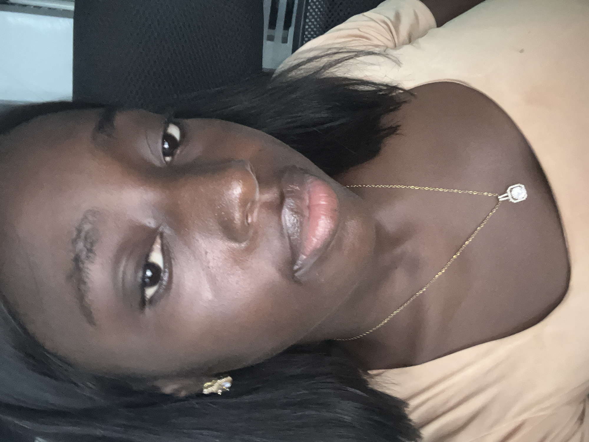 A dark-skinned woman is seen in a close-up, slightly angled shot. She is wearing a light-colored top and a gold necklace with a small, diamond-like pendant. She is also wearing an earring. Her dark hair is visible around her face and shoulders. The background is blurry and indistinct, but possibly includes a black office chair. There are no discernible features that would allow location in the city of Karum, Nigeria.