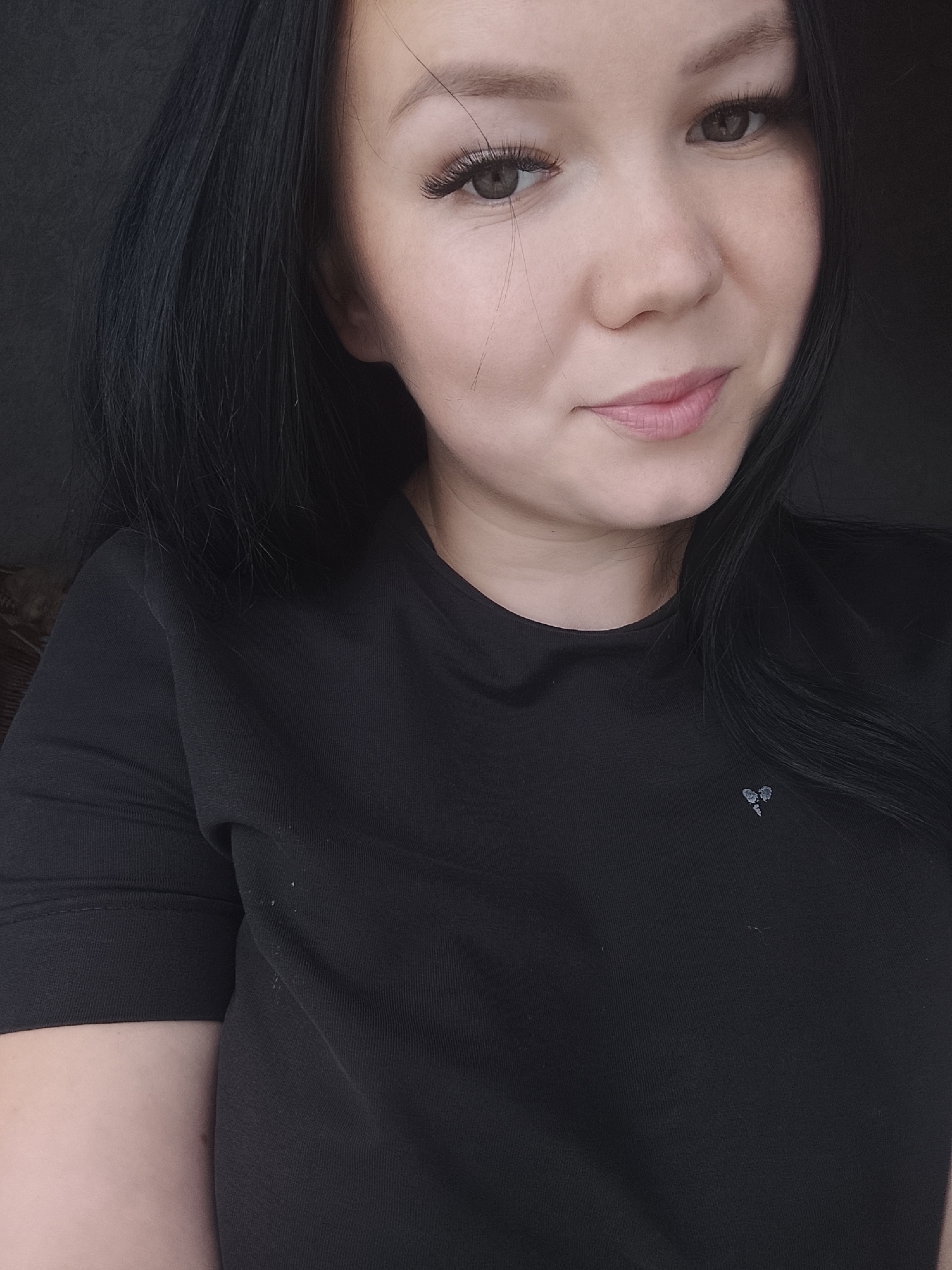 The image features a woman from the chest up, looking directly at the viewer with a slight smile. She has long, dark, straight hair, parted on the side and falling over her shoulders. Her complexion is fair, and she has brown eyes, enhanced with false eyelashes and eyeliner. Her lips have a light pink tint. She is wearing a dark, likely black, short-sleeved top or t-shirt made of a knit fabric. A small, greyish-white emblem, resembling two intertwined, somewhat fractured heart shapes, is visible on the left side of her chest. The background is dark and largely indistinct, suggesting an indoor setting with low light. A subtle textured pattern, possibly from furniture or a wall, is faintly visible on the left side of the frame. There are no discernible visual cues within the image to identify or confirm the specified location of Kuyeda, Russia.