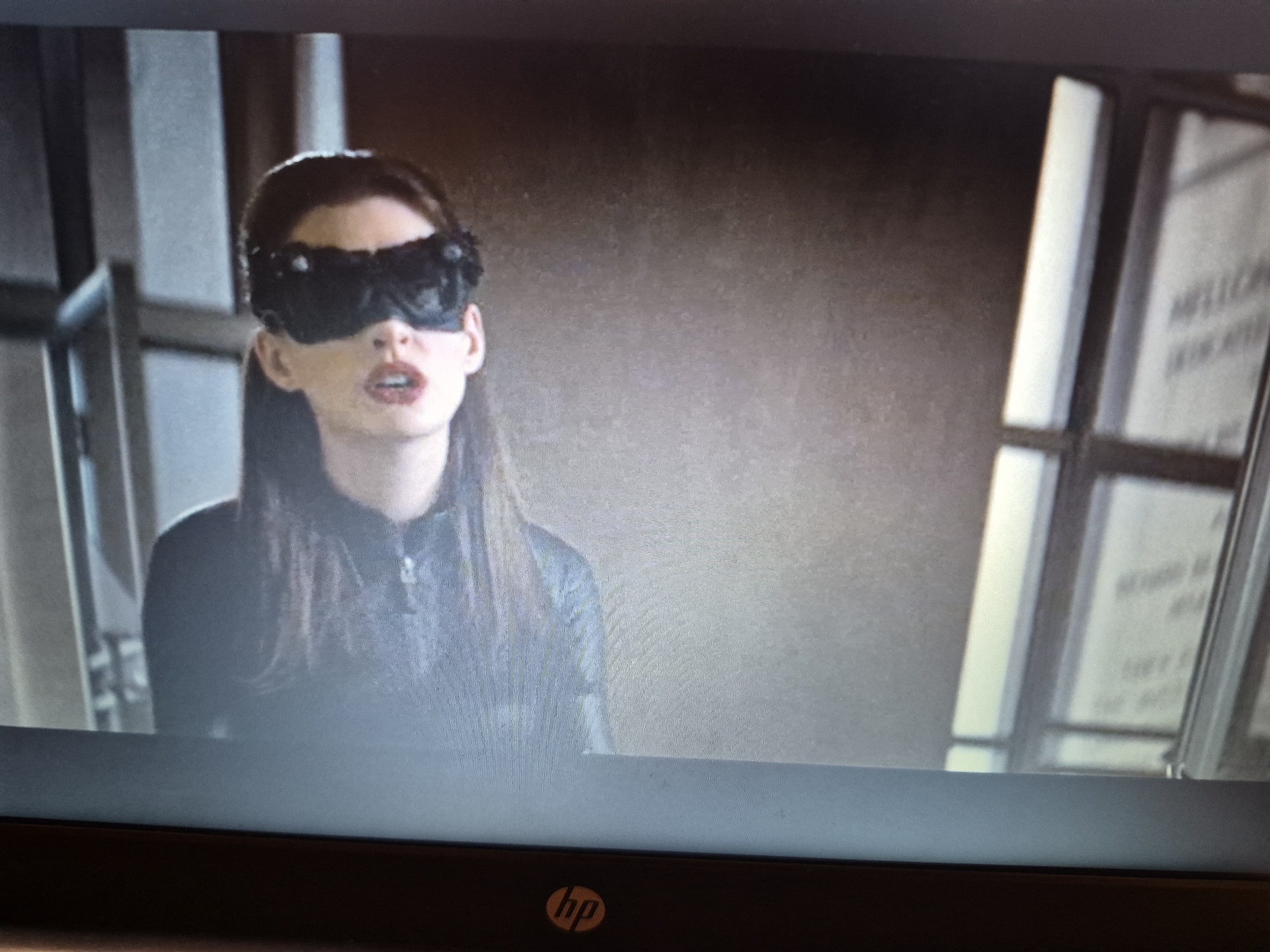 A close-up shot shows Anne Hathaway as Catwoman from the movie The Dark Knight Rises. She is wearing a black lace mask that covers her eyes and extends to her temples, with a dark, textured costume. Her mouth is slightly open, and her hair is dark and wavy, falling over her shoulders. The background is dimly lit and appears to be indoors, with vertical elements that could be part of a building's architecture or a set. The image is captured on a screen, indicated by the pixelated quality and the presence of an HP logo at the bottom. There is no discernible information within the image to suggest a location in the city of Sandwell, United Kingdom.