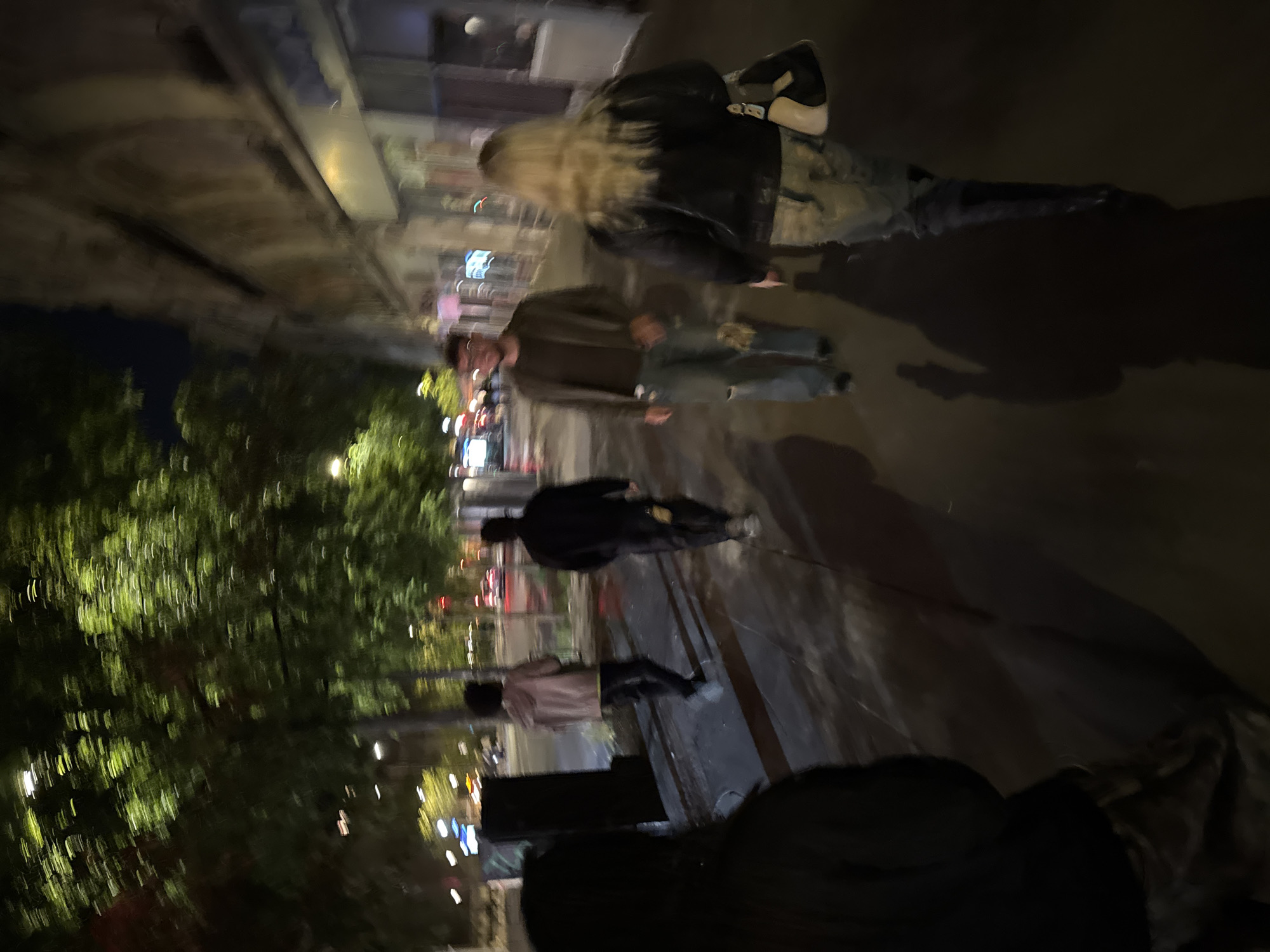 A blurred, low-angle shot depicts a nighttime urban scene, likely in Paris, France, suggested by the architectural styles and lighting. Four pedestrians are visible on a dimly lit sidewalk, their images distorted by motion blur. To the top right of the frame, two people walk in the same direction. The person closest to the camera has long, blonde hair and is wearing a black jacket, jeans, and white shoes. The person beside them is wearing glasses, a brown shirt, and blue jeans. Down the sidewalk, two more people are visible, with one person wearing a black jacket, and the other a pink shirt. To the left of the scene, there are trees.