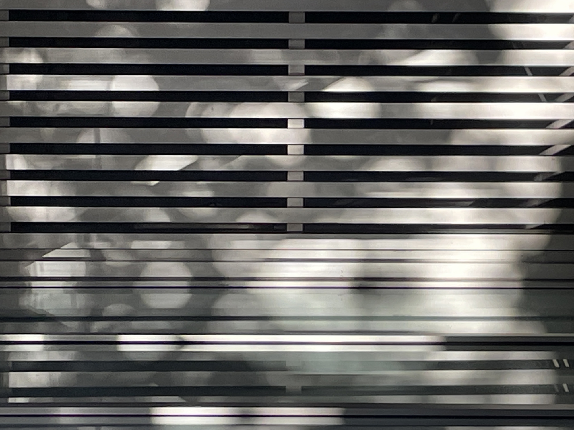 The image features an abstract architectural detail, likely a building facade, louvered window, or industrial shutter, in Buenos Aires, Argentina. The main subject is a grid-like pattern of alternating light gray and dark gray or black horizontal strips, creating a strong sense of rhythm and depth. These strips are uniformly spaced across the frame.
Overlaid on this structured background is a dynamic interplay of light and shadow. Bright, diffused light spills across various sections, highlighting the reflective quality of the lighter strips. Irregular, organic shadow patterns are cast onto the surface, suggesting that sunlight is filtering through or reflecting off nearby foliage, structures, or other elements. These shadows are soft-edged and vary in intensity, creating a sense of movement and depth. Several blurred, bright circular or amorphous shapes, similar to bokeh, are visible within the illuminated areas, indicating out-of-focus light sources or reflections.
The strong contrast between the bright and shaded areas, along with the distinct patterns of light, suggests a bright, sunny daytime setting. The image is devoid of people or specific identifiable objects beyond the architectural elements and the play of light. No visible text is present. The overall composition is a study in light, shadow, and texture on a repetitive, man-made surface.