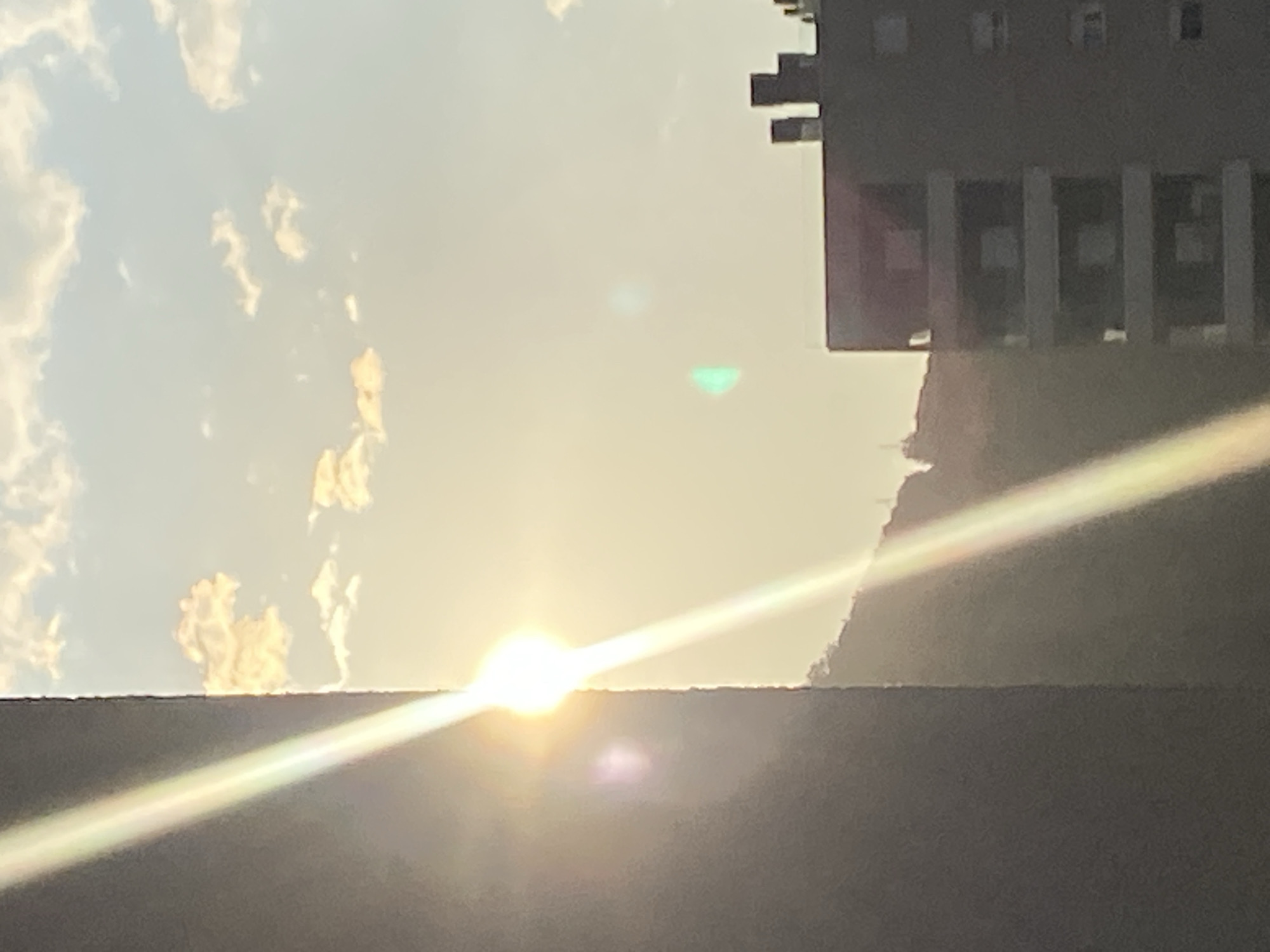 The image shows a bright sun obscured by a dark, horizontal structure, possibly a building or wall. Above the sun, there are light-colored clouds against a blue sky. To the right of the sun, a darker mountain slope and a building with visible columns and windows are present. The bright light of the sun causes lens flare. The location is likely Budva, Montenegro, given the architecture and mountainous terrain.