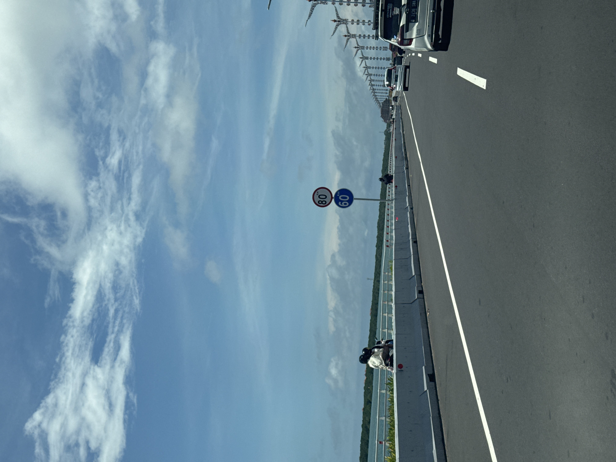 The image shows a section of the Bali Mandara Toll Road in Nusa Dua, Indonesia. The road is a gray asphalt surface with white lane markings, currently bearing moderate vehicular traffic including cars. A white barrier is on the left side of the road, featuring decorative red reflectors. Speed limit signs are present, reading "80" and "60". The background reveals a body of water, most likely a lagoon or bay, and lush vegetation on the far shore. The sky is blue with scattered white clouds.